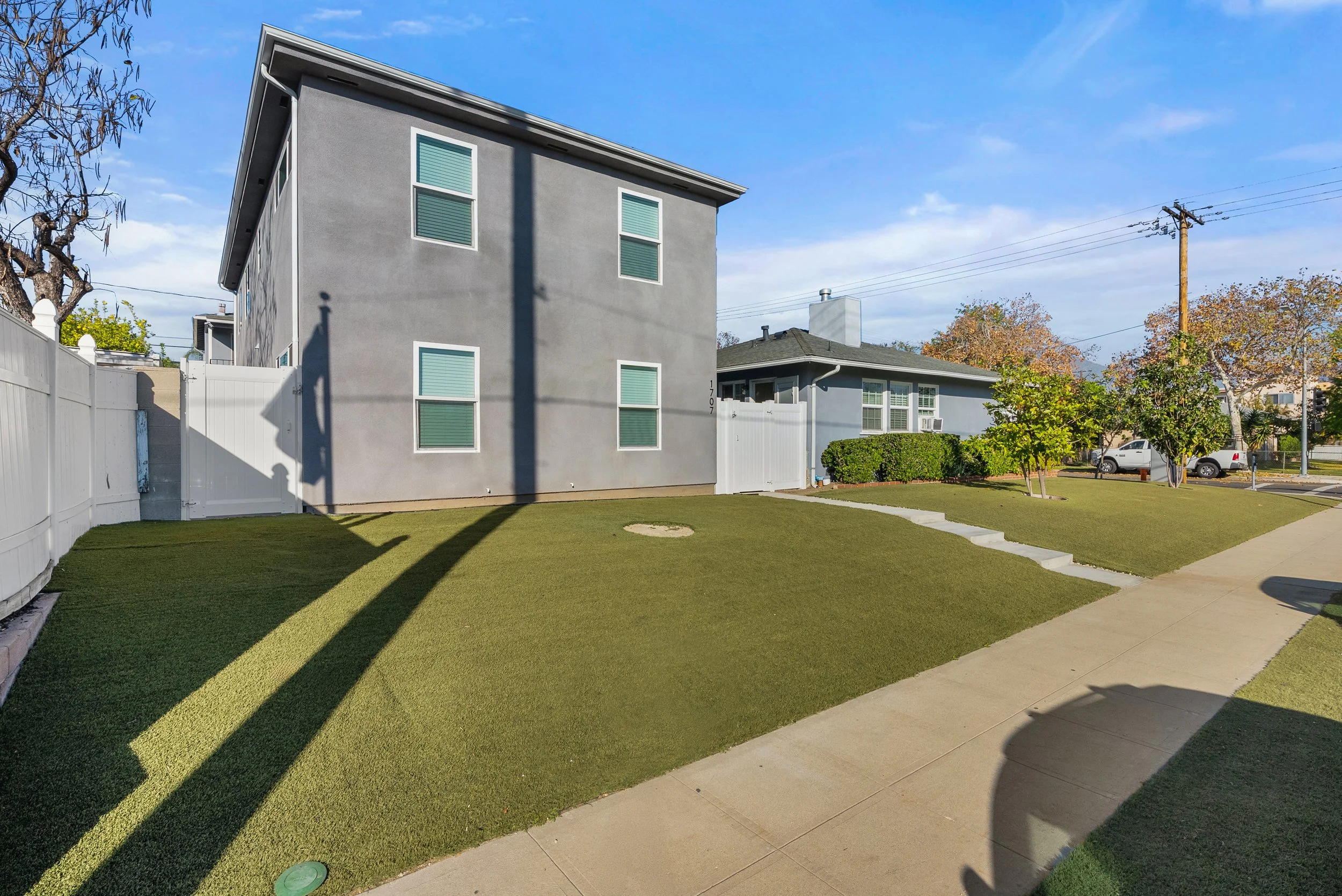 A residential house with gray exterior walls, four windows, a white fence, a small grass lawn, a concrete sidewalk, and trees, under a blue sky with some clouds.