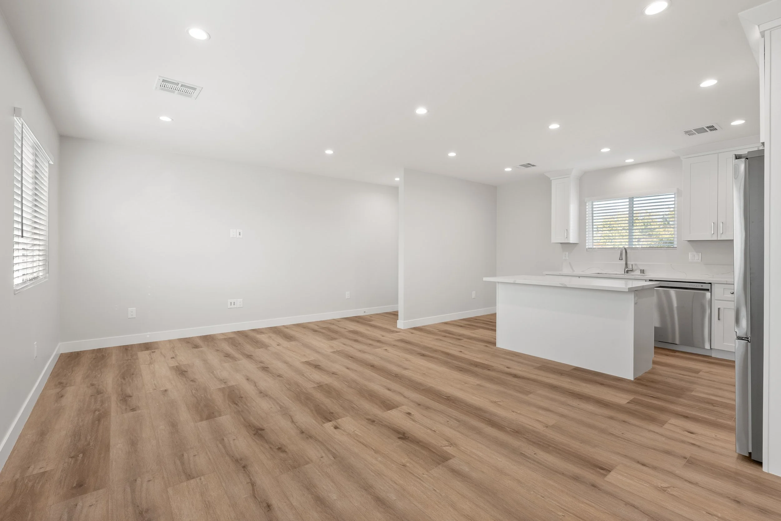 Empty open-concept living area with white walls, hardwood floors, and modern kitchen with white cabinets, a window, and stainless steel appliances.