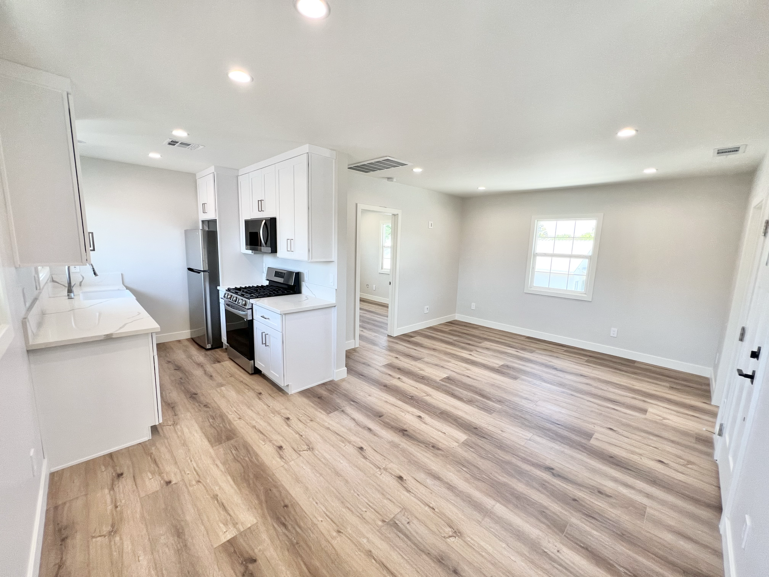 Empty open-concept kitchen and living room with white cabinets, stainless steel appliances, light wood flooring, and a large window.
