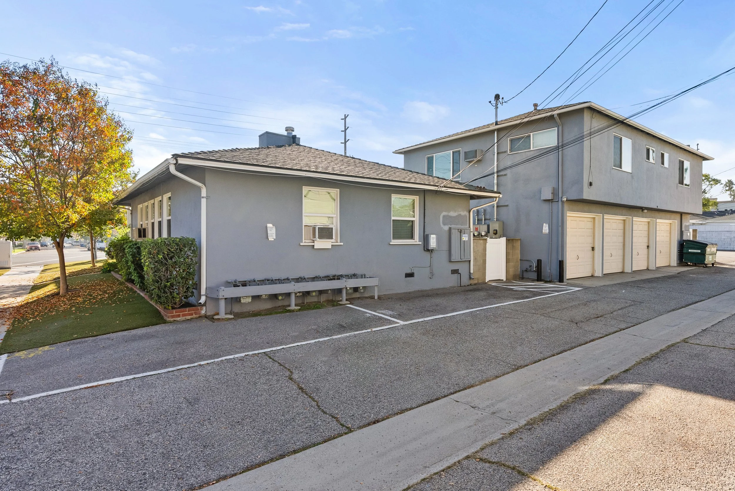 A two-story gray apartment building with a garage and parking lot in front, surrounded by a few trees with fall-colored leaves, under a blue sky.