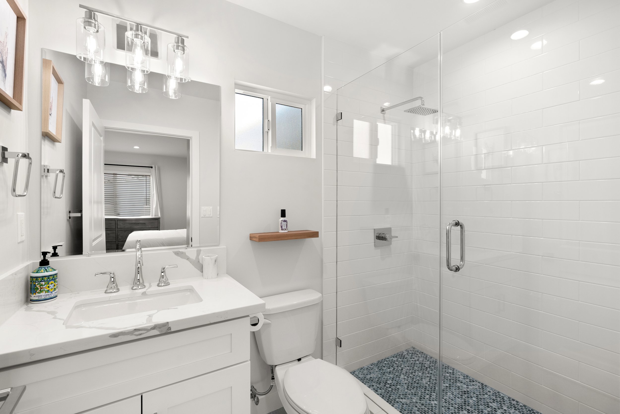 A modern, white bathroom with a vanity, mirror, small shelf, and a walk-in shower with a glass door and tiled shower floor.