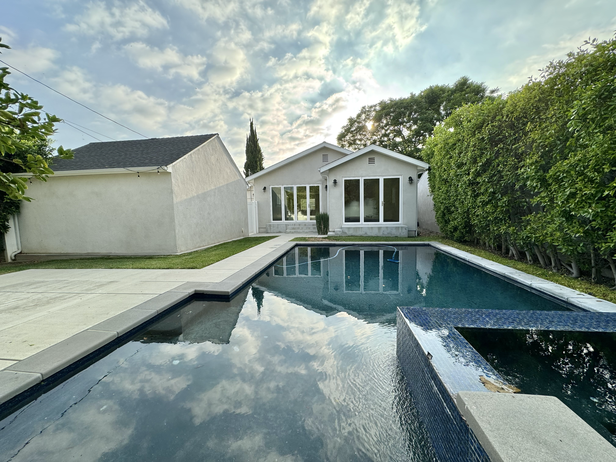A backyard with a swimming pool and a white house with sliding glass doors, surrounded by green bushes and a partly cloudy sky.