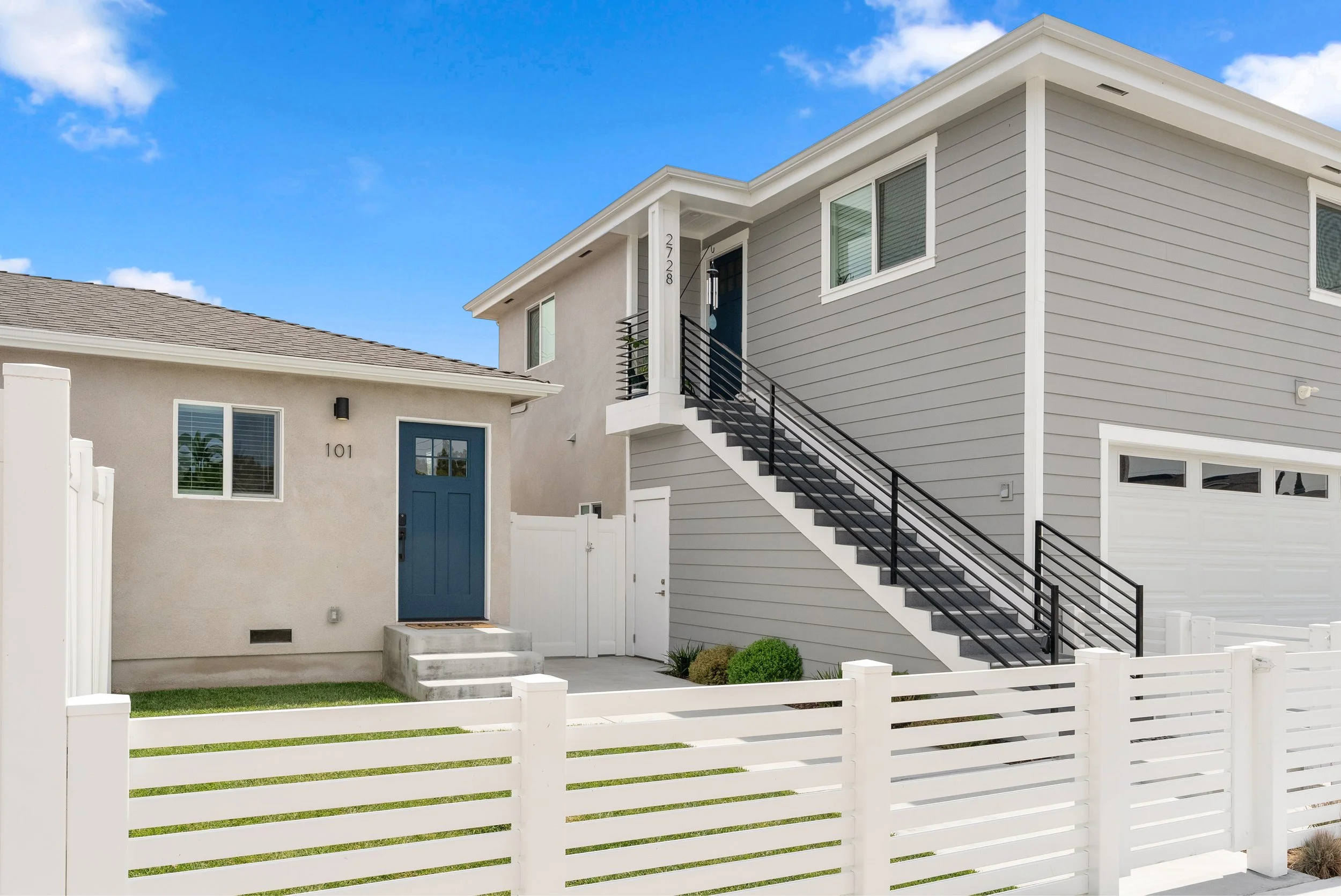 Modern two-story gray house with a front staircase, white fence, and blue front door, under a partly cloudy sky.