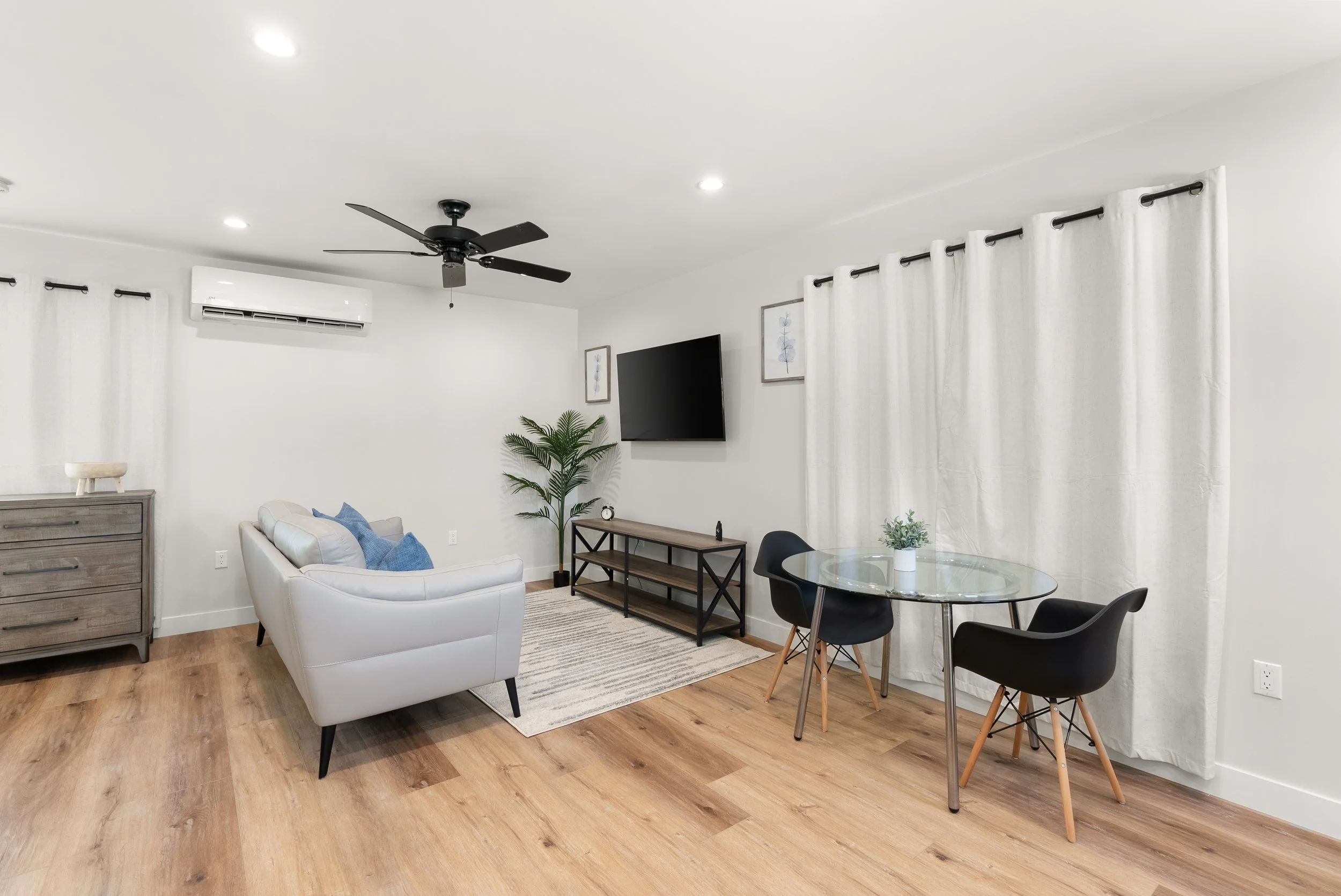 Living room with white walls, hardwood floors, a white sofa with blue pillows, a gray dresser, a large potted plant, a wall-mounted TV, a black ceiling fan, white curtains, and a glass dining table with three black chairs and a small potted plant.