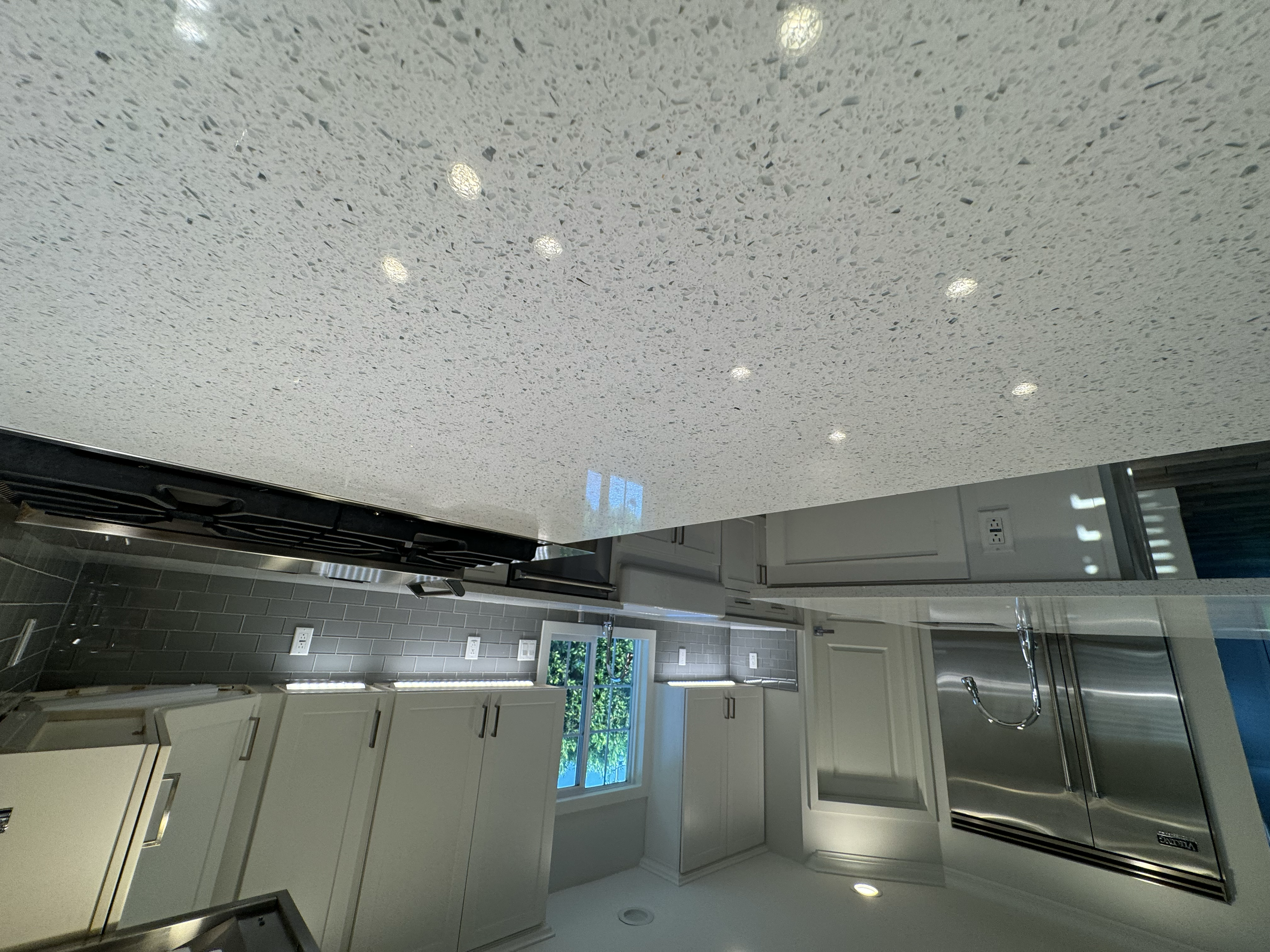 A modern kitchen with white cabinets, stainless steel appliances, and a gray subway tile backsplash. There are multiple electrical outlets and a window with greenery outside.