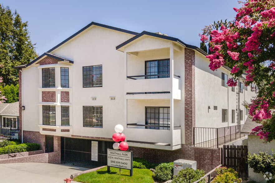 Multi-story white residential building with brick accents, surrounded by greenery and a flowering pink tree.