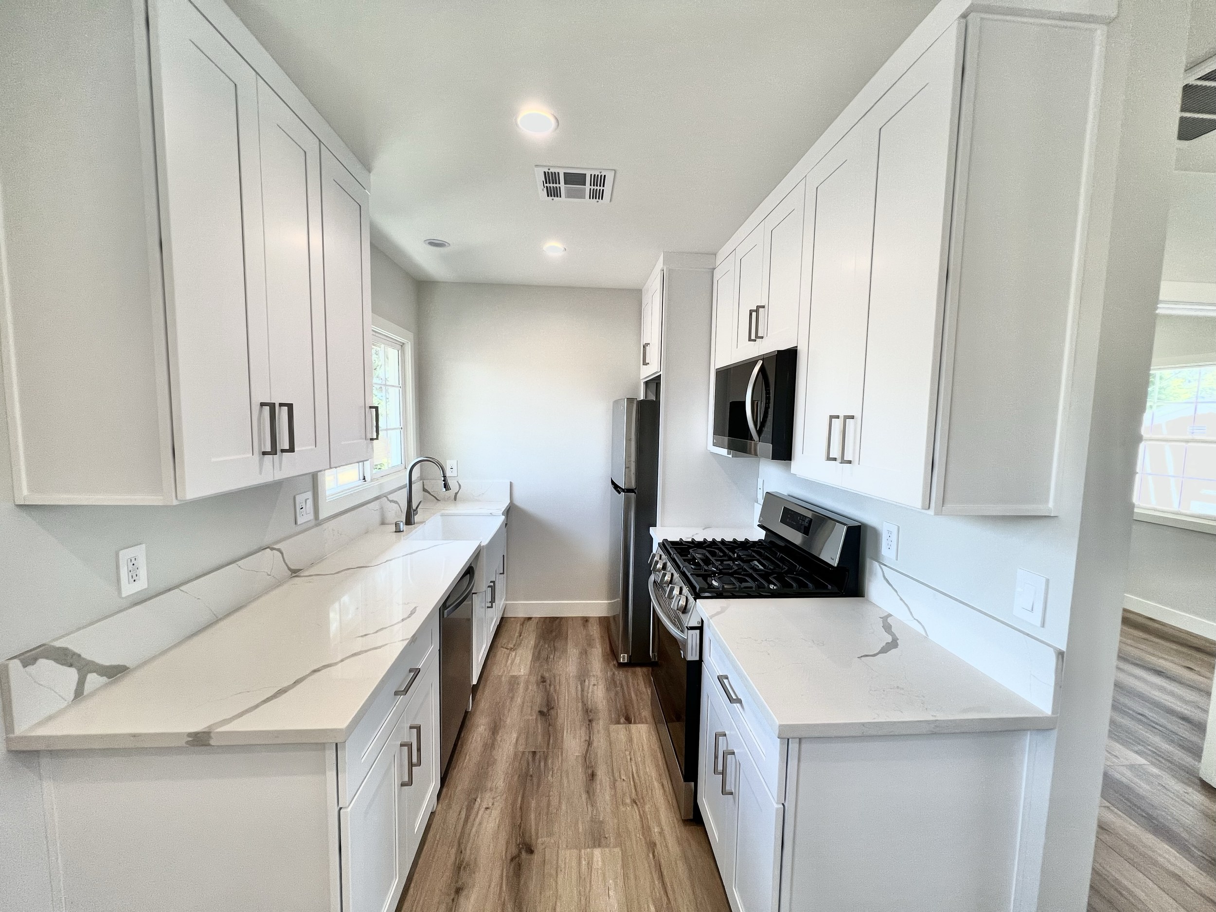 Modern white kitchen with marble countertops, stainless steel appliances including a refrigerator and stove, white cabinets, wood flooring, and a window above the sink.