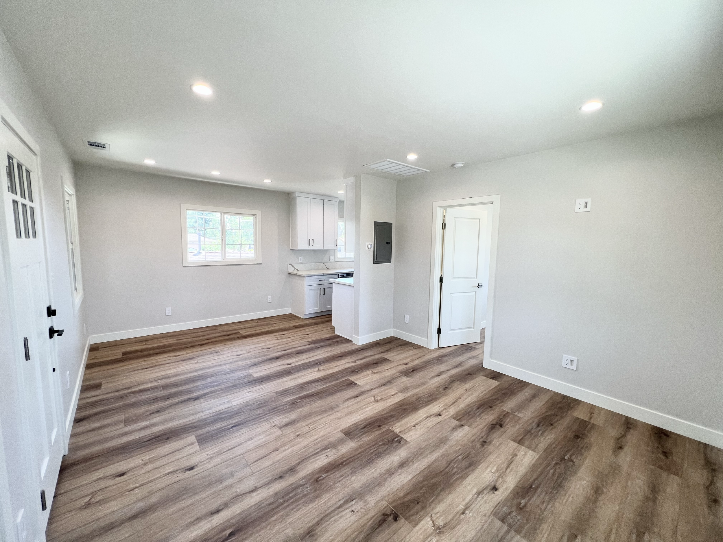 Empty living space with wood flooring, white walls, recessed ceiling lights, a window, and an open kitchen area.
