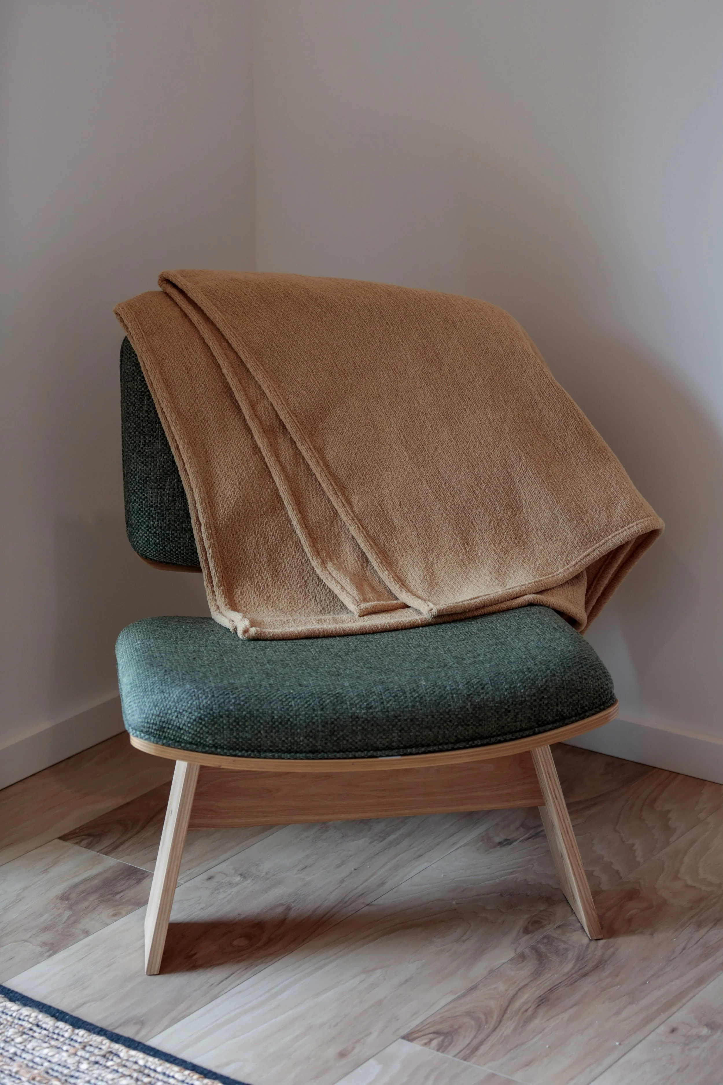 A mid-century modern style green upholstered lounge chair with light wood legs, positioned in a corner with a plain off-white wall background, a beige throw blanket draped over the backrest, and a patterned rug partially visible at the bottom of the image.