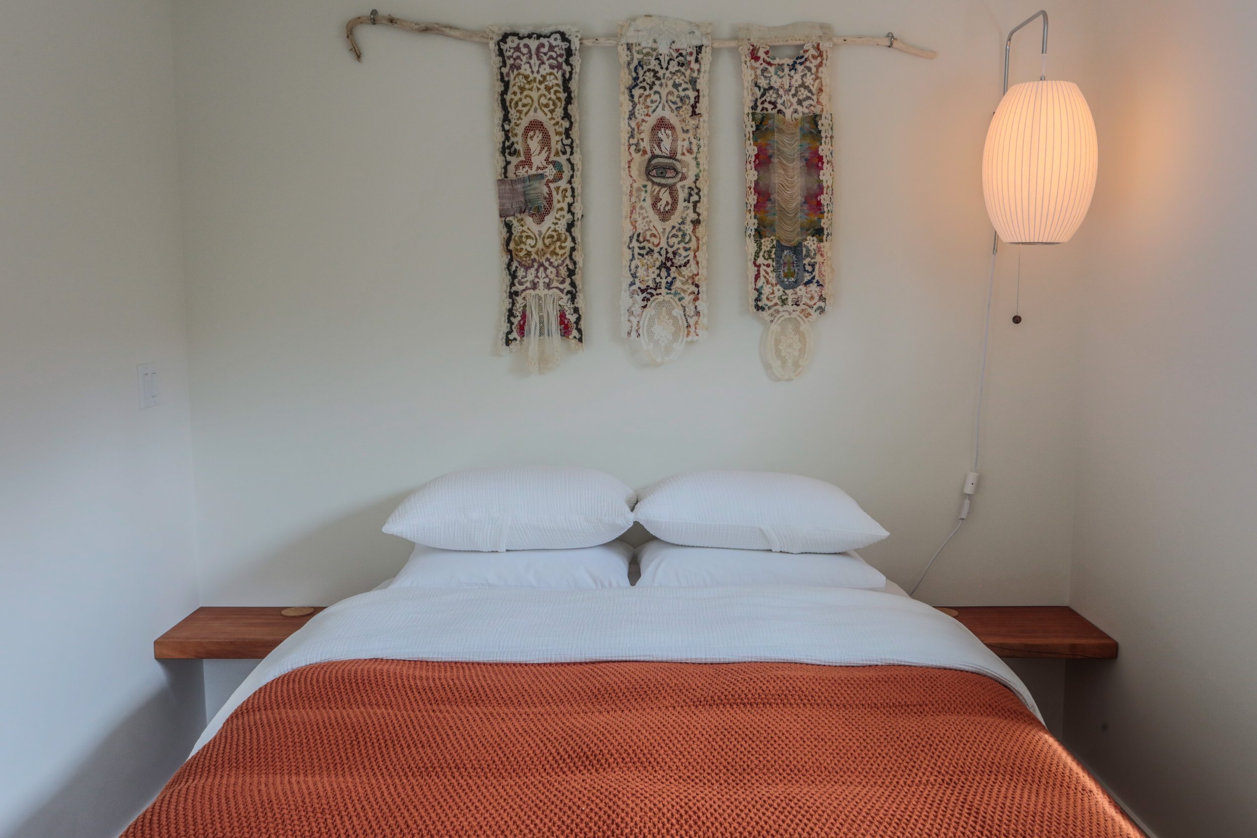 A bed with white linens and two pillows, with a wooden headboard and an orange blanket at the foot. Above the bed, there is a decorative wall hanging made of woven fabric strips and a horizontal branch, next to a wall-mounted lamp with a paper shade.