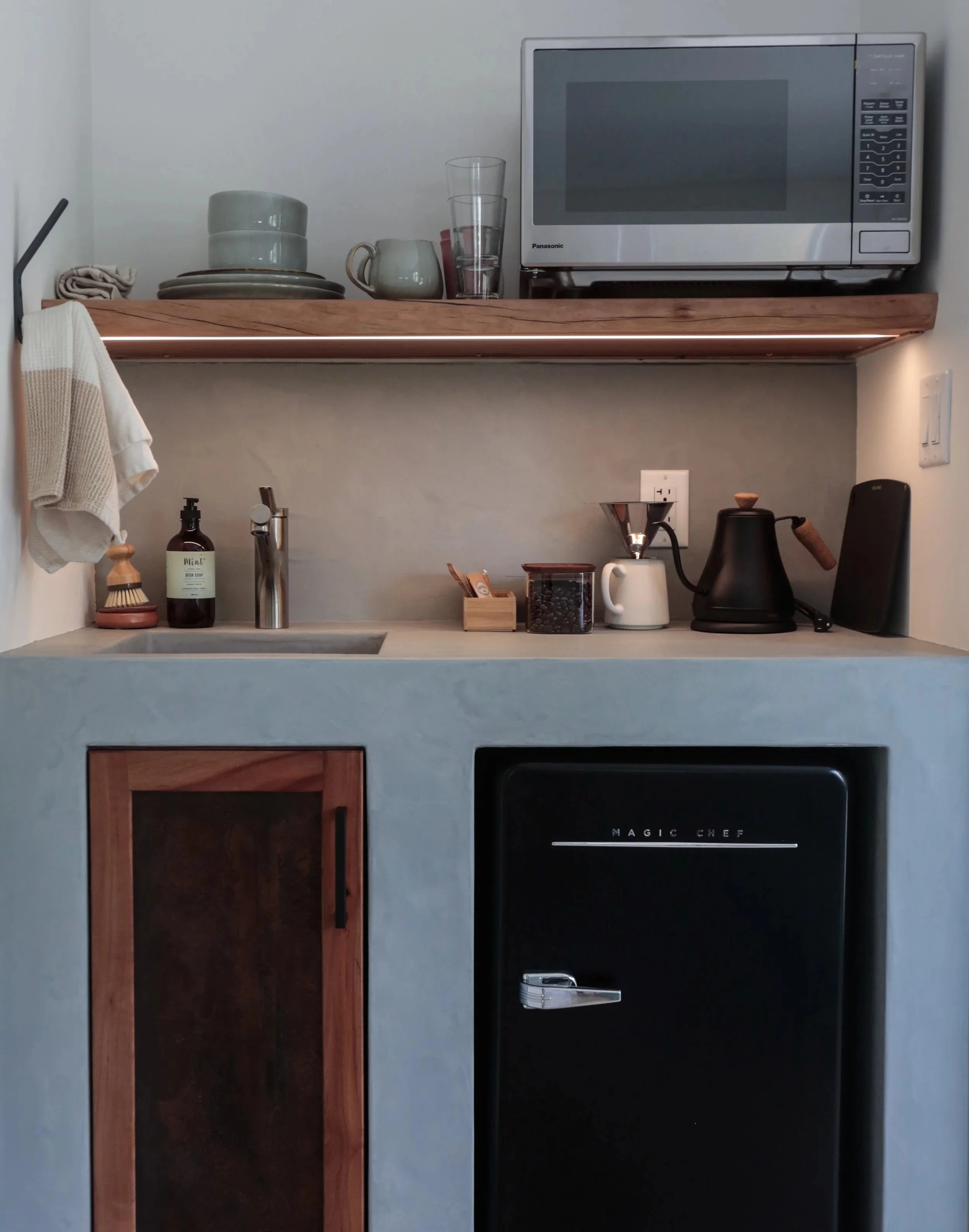 Kitchen counter with a microwave oven on the top shelf, a black mini fridge below, and various kitchen utensils and coffee-making items on the counter.