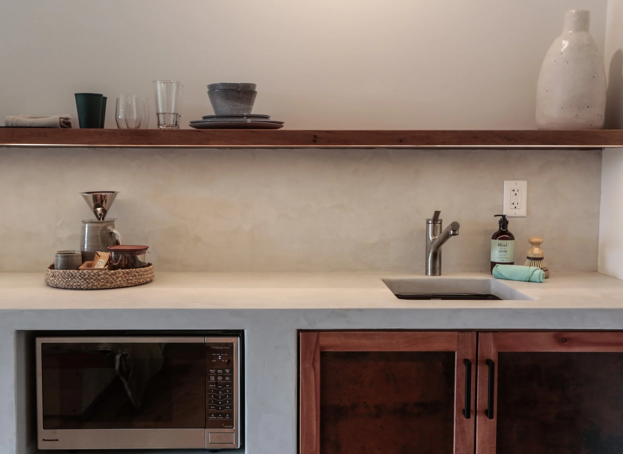 Kitchen counter with a microwave oven, a glass vessel with a pour-over coffee brewer, a woven basket with small containers, and a bottle of dish soap.