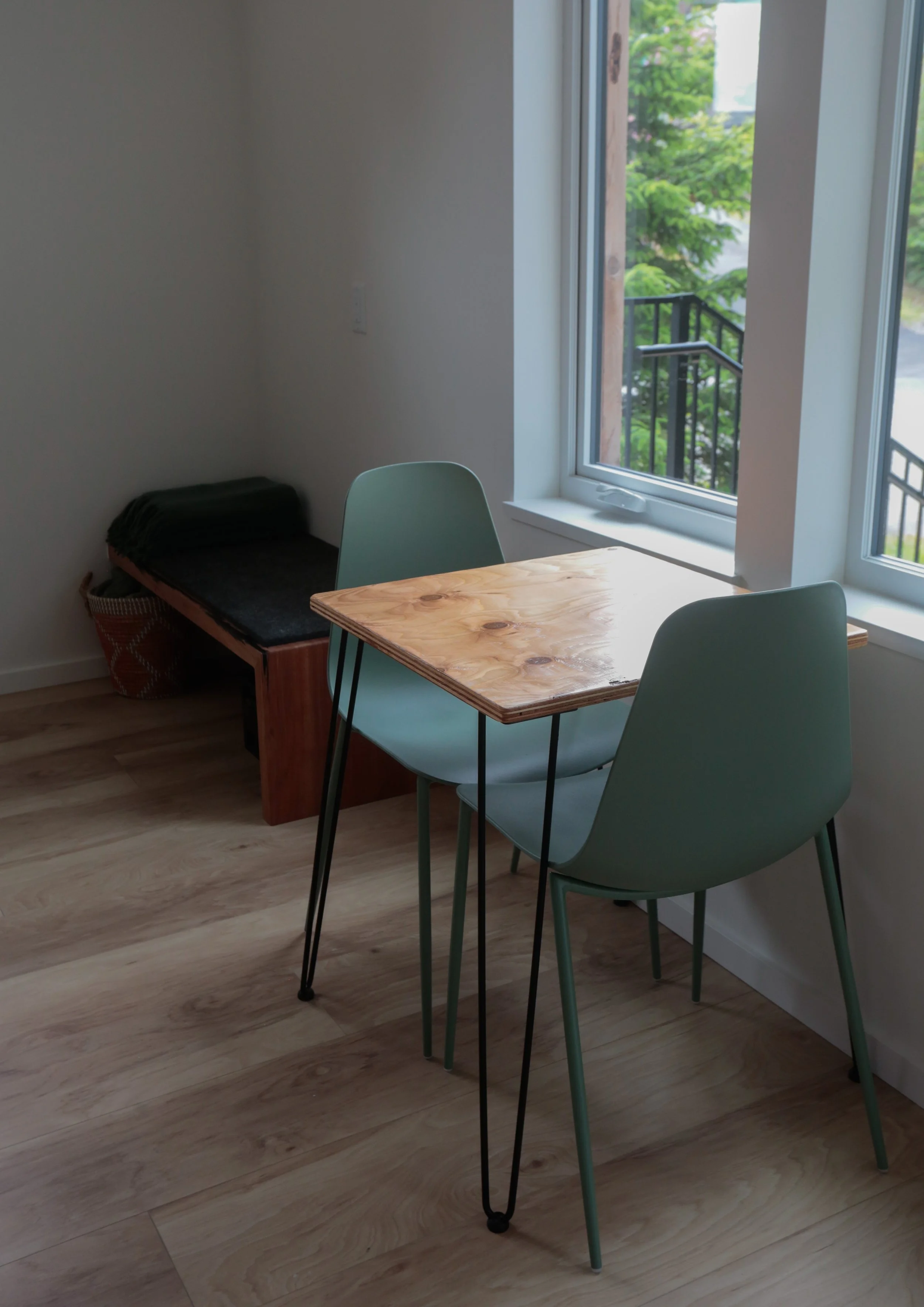 A cozy interior space with a wooden table, two green chairs, a small black bed, and a large window with a view of green trees outside.