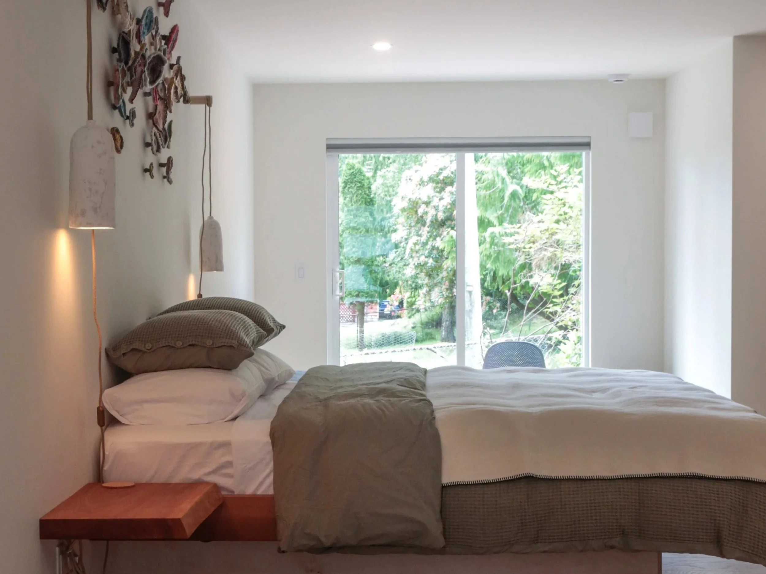 A bedroom with a bed that has multiple pillows, a side table, a hanging lamp, and a large window showing greenery outside.