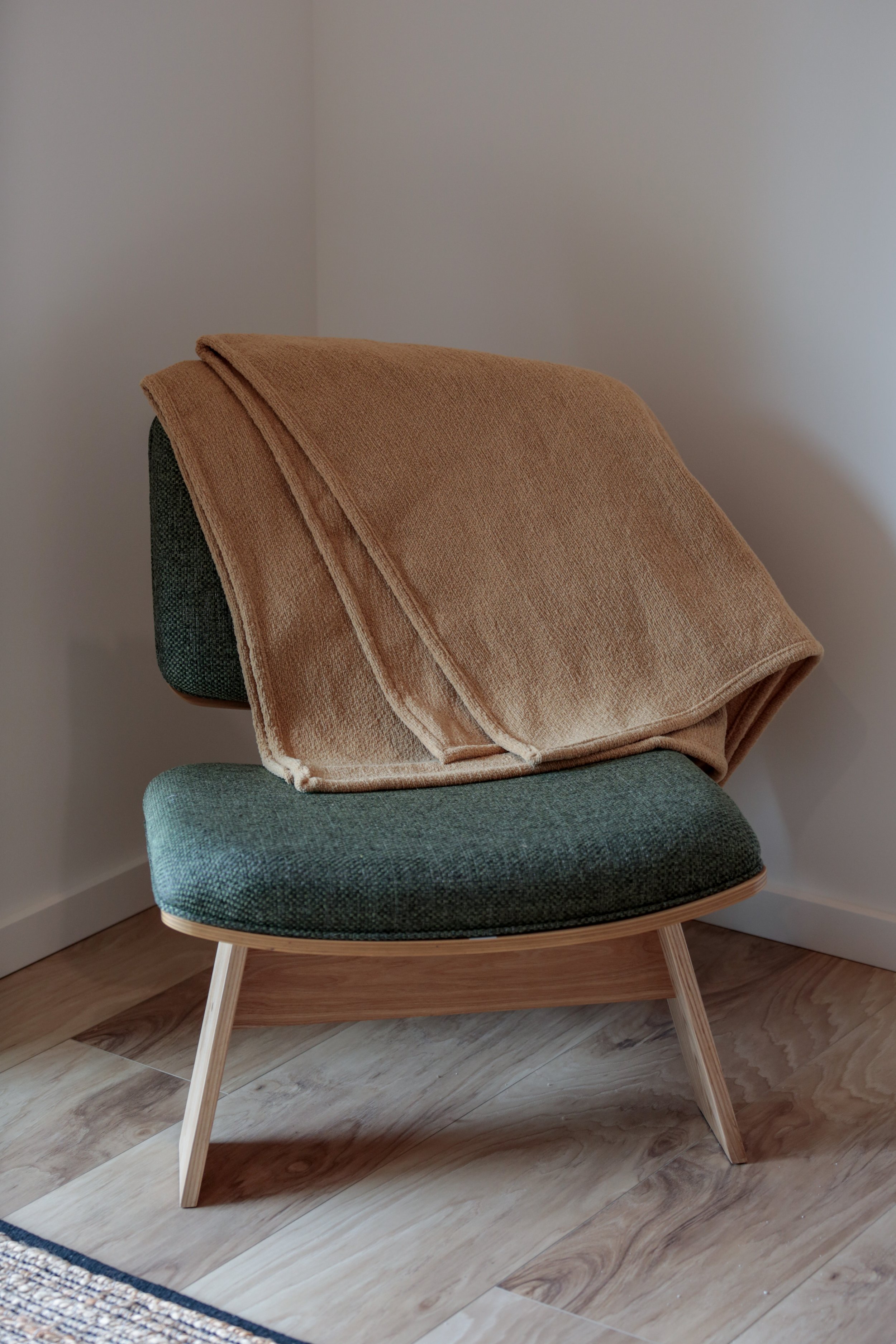 A modern green upholstered armchair with wooden legs, placed near a corner wall with a beige blanket draped over its back.