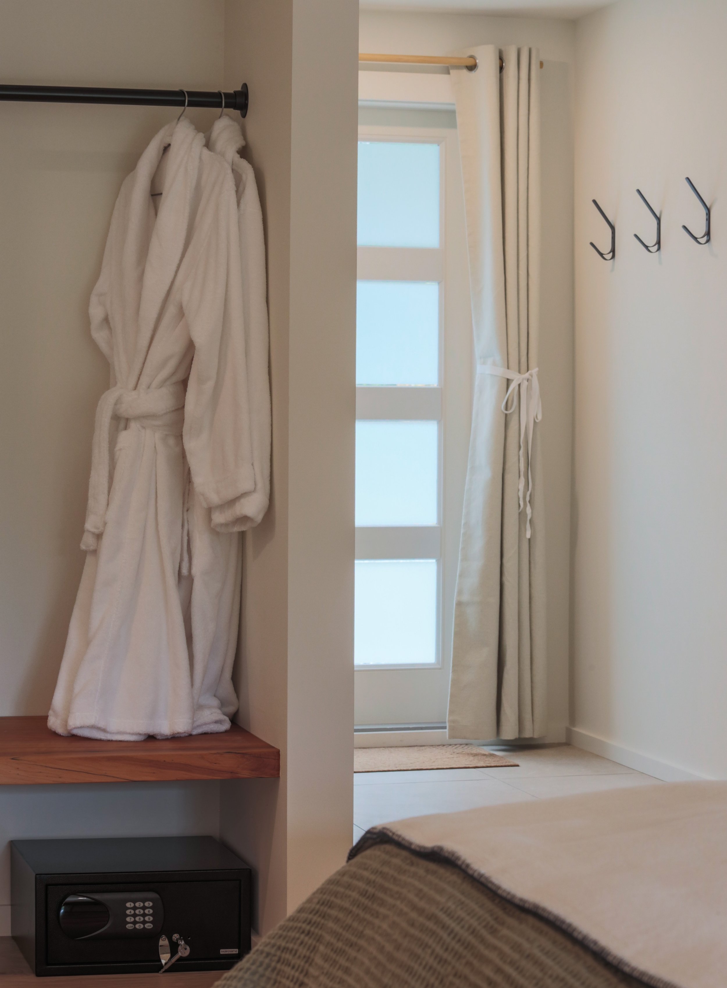 A view from a bedroom showing a closet with white bathrobes hanging on a black rod, a window with white curtains, and a safe below the closet.