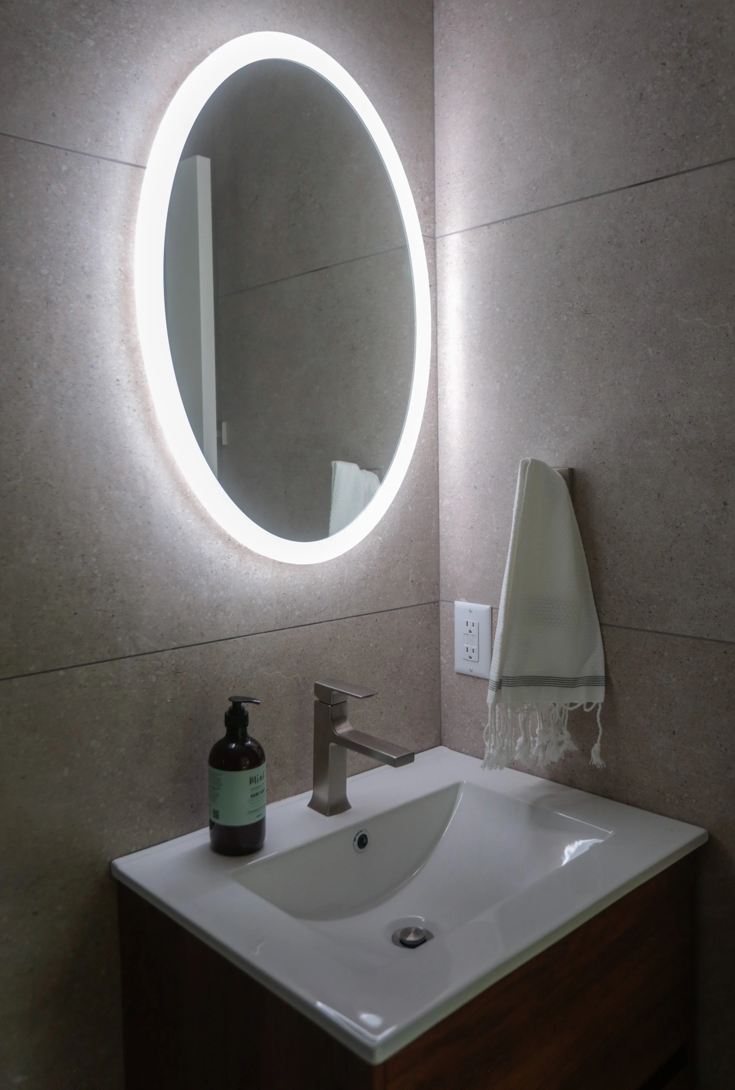 Bathroom sink with a silver faucet, a bottle of hand soap, a round mirror with LED lighting, a white towel, and a beige tiled wall.