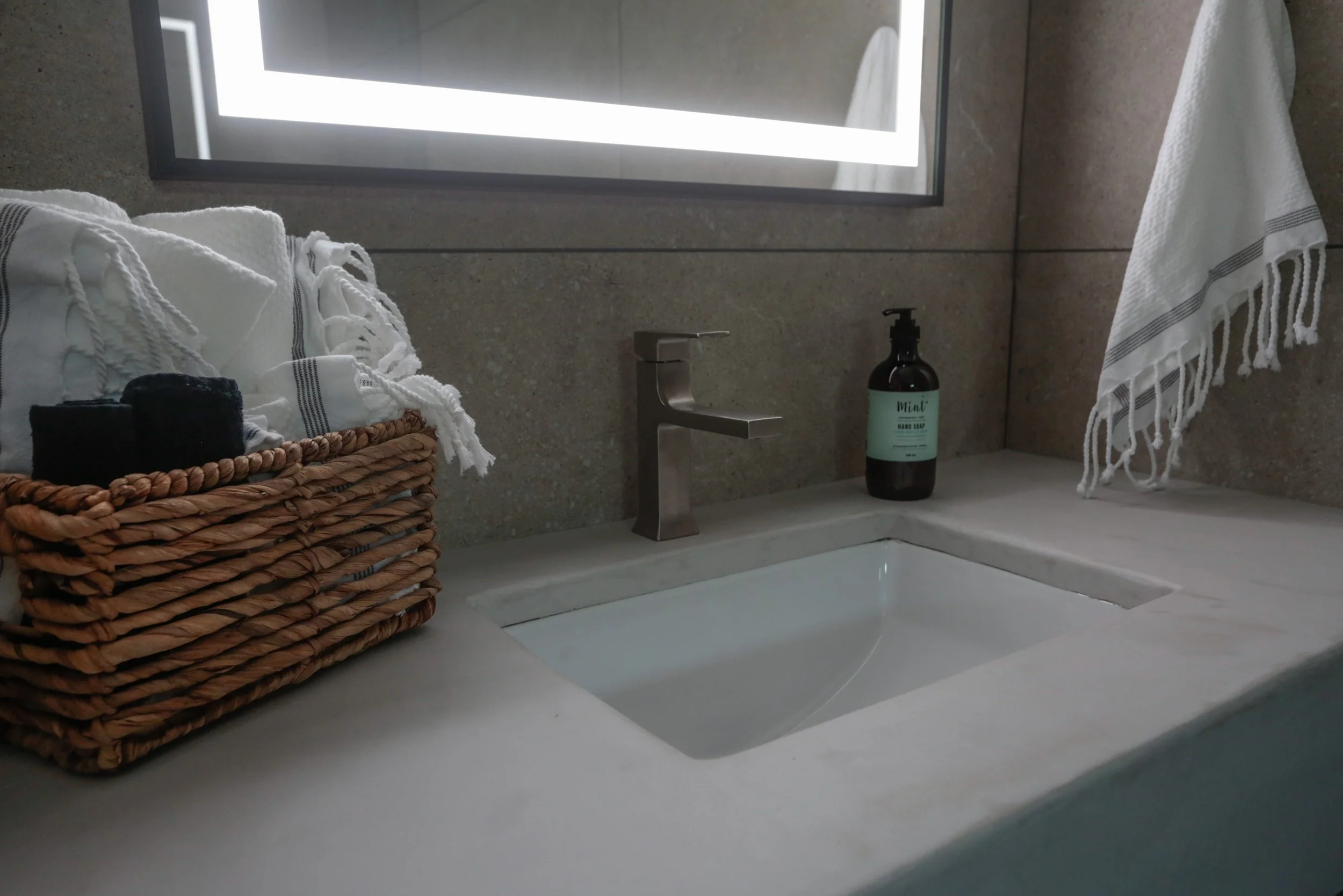 Close-up of a bathroom sink with a woven basket of rolled towels, a soap dispenser labeled 'Mint', a silver faucet, and a white towel with fringes hanging on the side. Mirror and wall behind the sink.