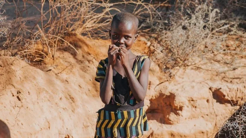 A young girl with a shaved head and dark skin standing outdoors in a dry, rocky area with sparse bushes, wearing a colorful striped dress and smiling softly.
