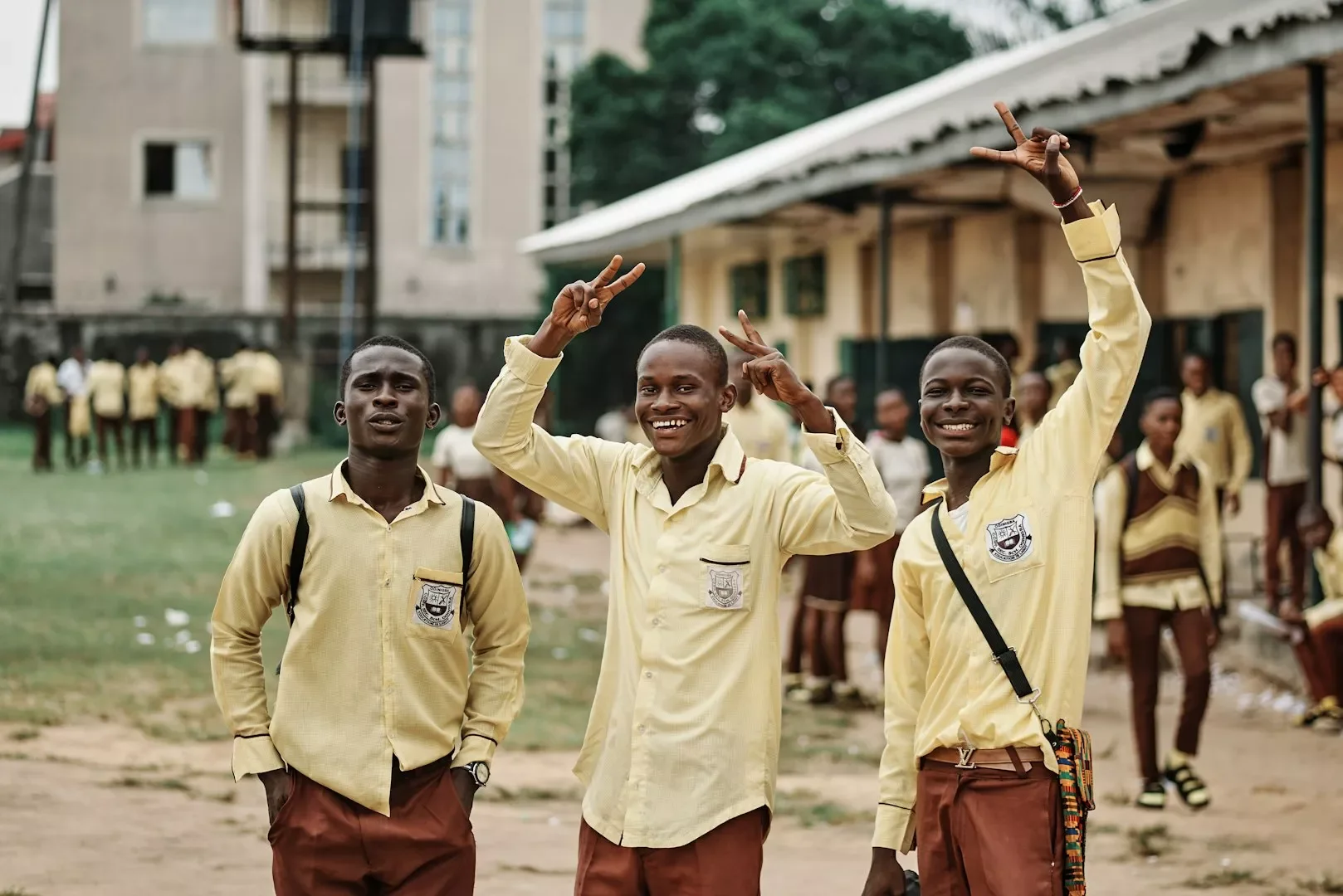 Three young boys in yellow school uniforms smiling and making peace signs with their hands on a school playground, with more students in the background.