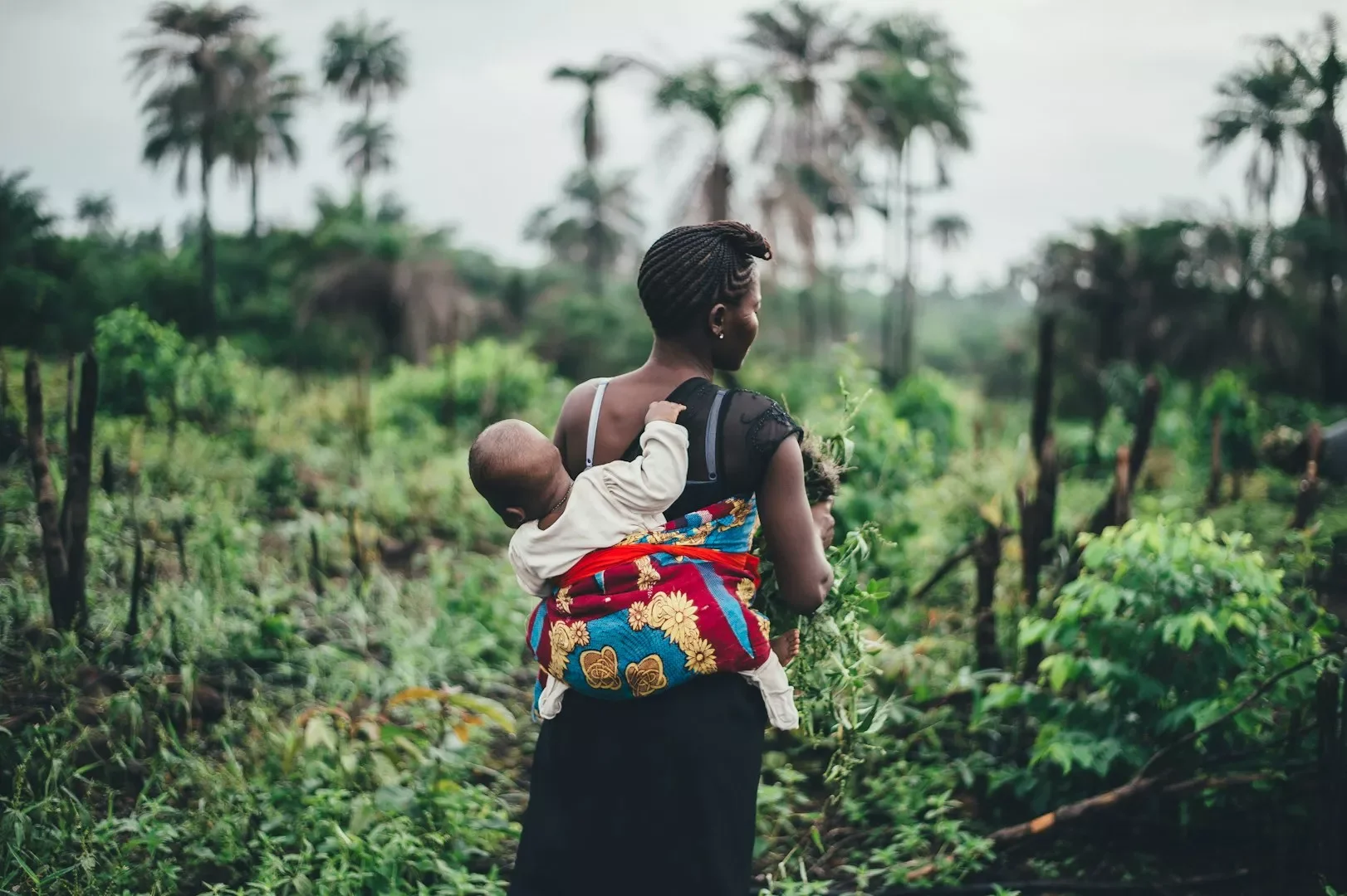 A woman walking through a lush, green landscape carrying a baby on her back and a young child holding onto her shoulder.