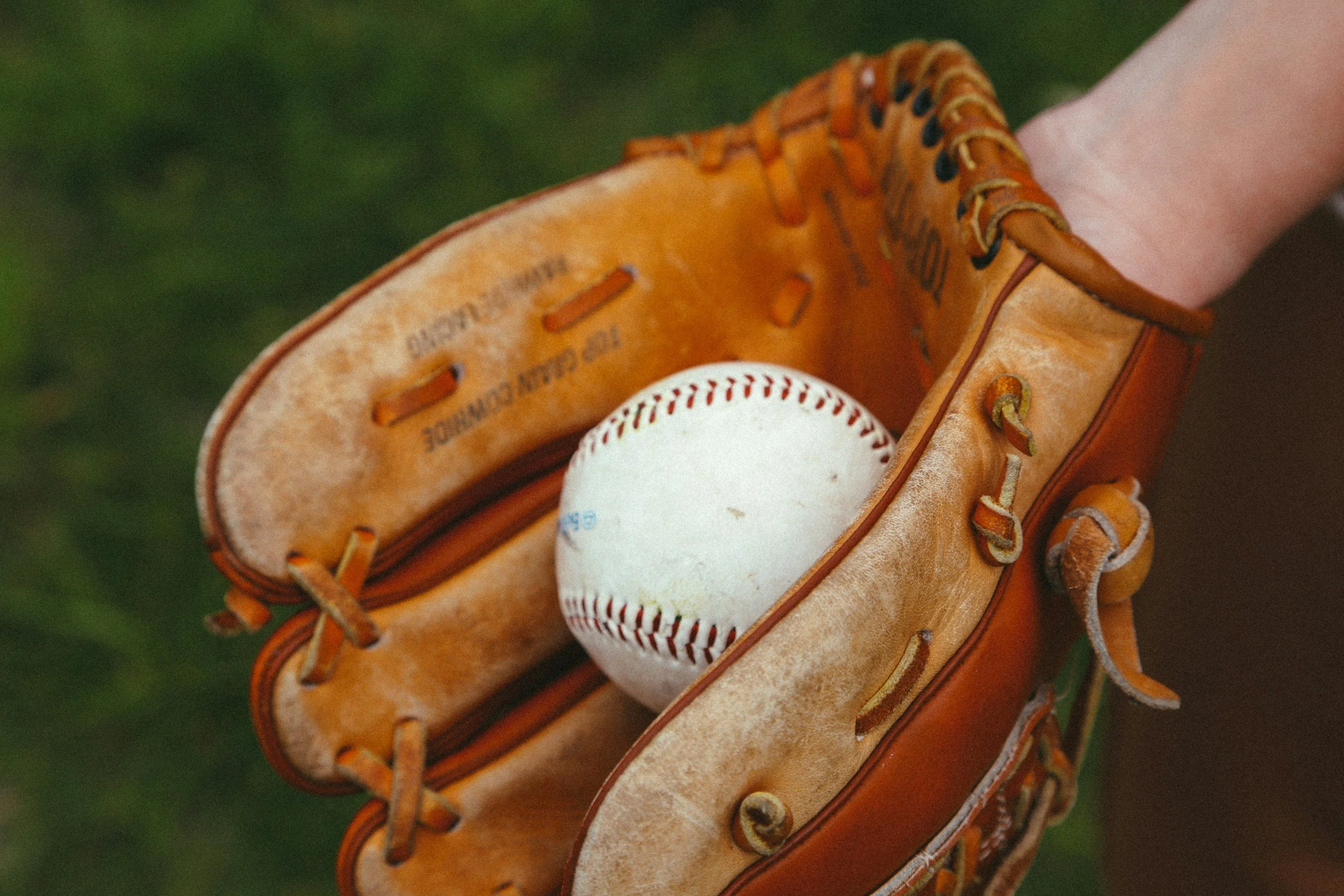 A leather baseball glove holding a white baseball, with a grassy background.