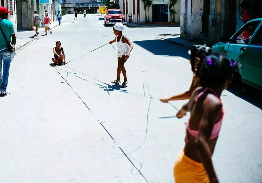 Children playing jump rope on a sunny street with cars and buildings in the background.