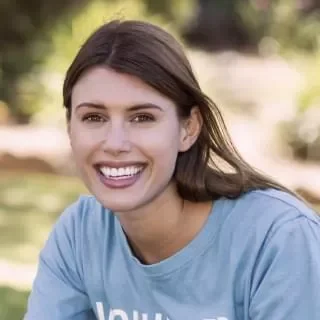 A smiling woman with long brown hair wearing a light blue shirt outdoors