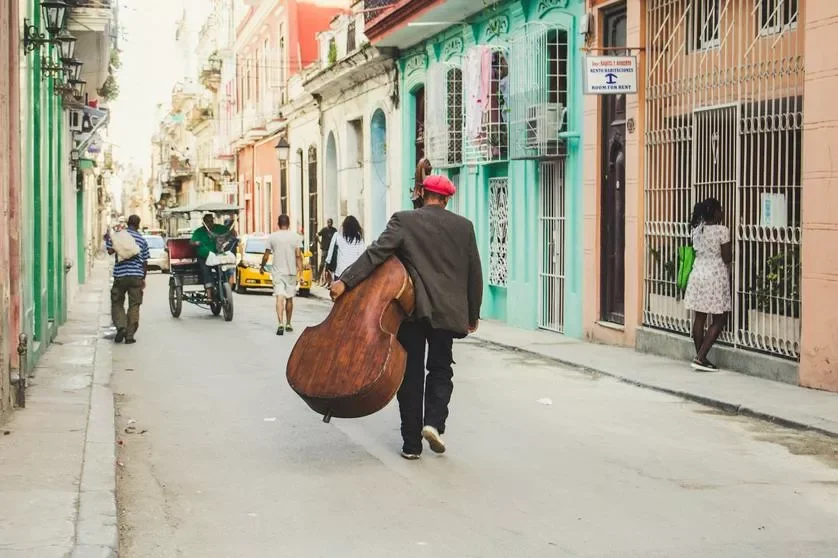 A man carrying a large bass guitar walking down a colorful city street with pedestrians and vehicles.