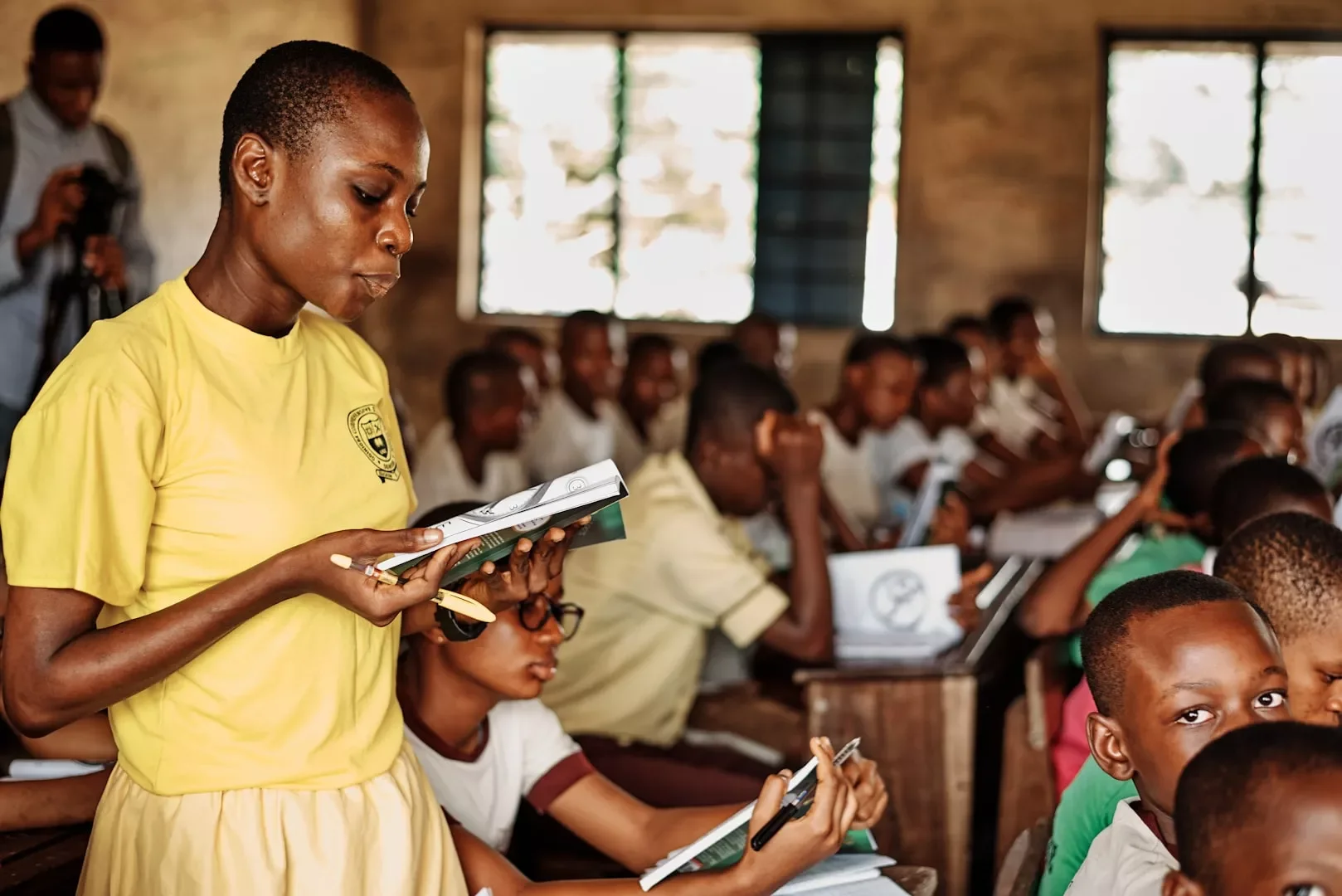 A classroom full of students, with a girl in a yellow shirt standing and reading a notebook while others are seated, some using laptops or taking notes, during daytime.