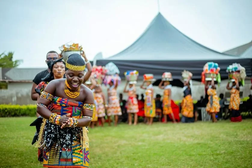 African woman smiling and dancing in traditional attire with other women carrying baskets on their heads in the background at an outdoor cultural event.