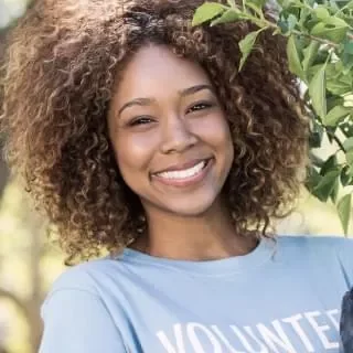 A young woman with curly hair smiling outdoors in front of green leaves, wearing a blue t-shirt.
