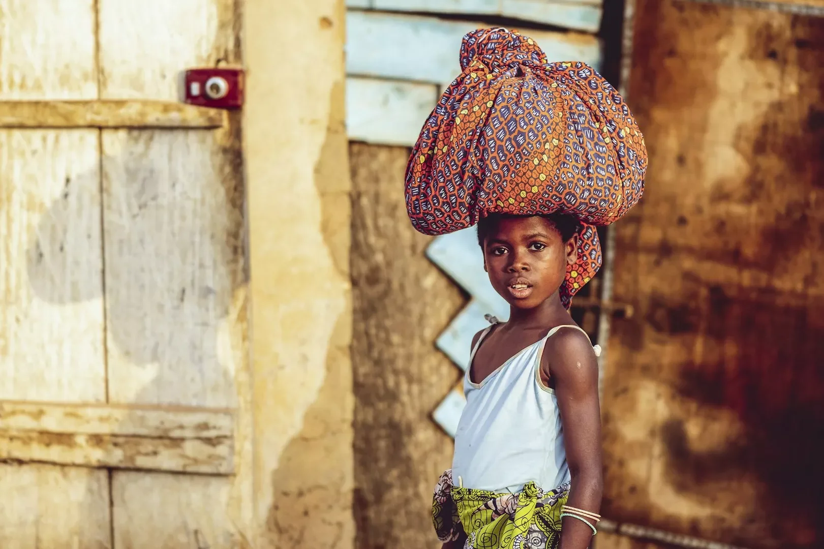 A young girl with dark skin, wearing a light blue sleeveless dress, stands outdoors holding a large colorful fabric bundle on her head. The background features a rustic building with wooden and mud walls.