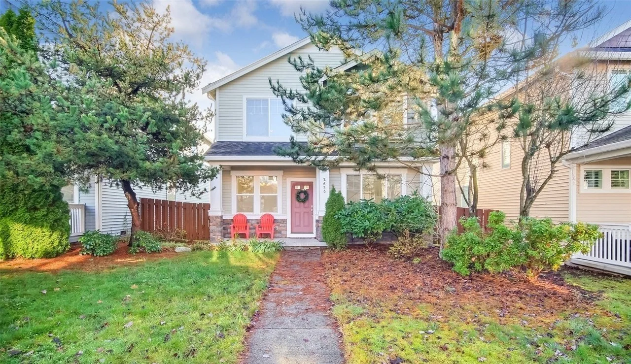 Front view of a suburban house with two stories, trees, and shrubs, featuring a walkway, two red chairs, and a wreath on the door.