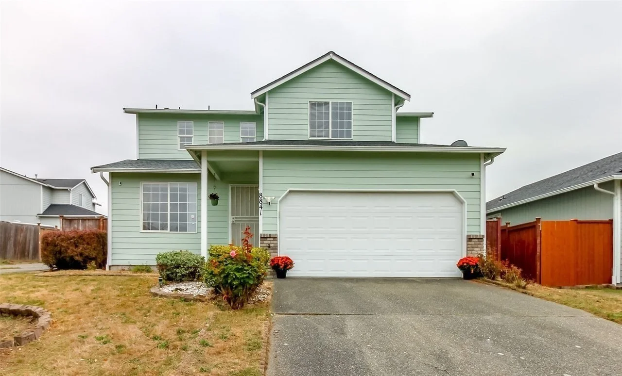 Two-story light green house with a white garage door, front lawn, and plants near the entrance.
