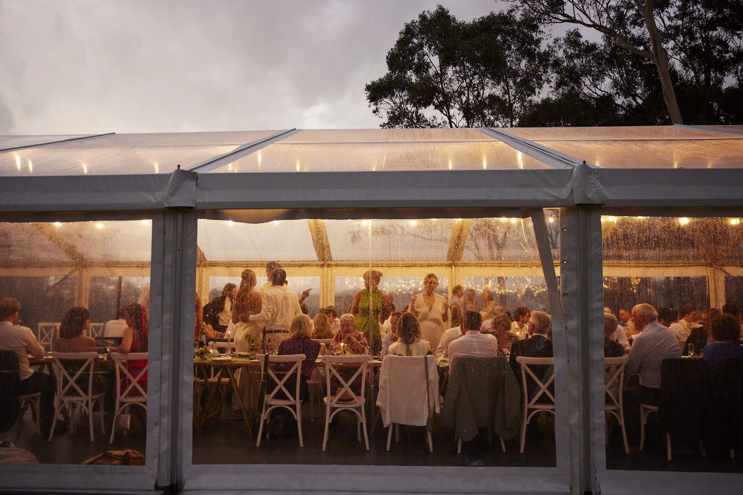 A large outdoor wedding reception at dusk, under a clear tent with string lights, where numerous guests are seated at tables and standing, at Dryridge Estate, Megalong Valley, NSW