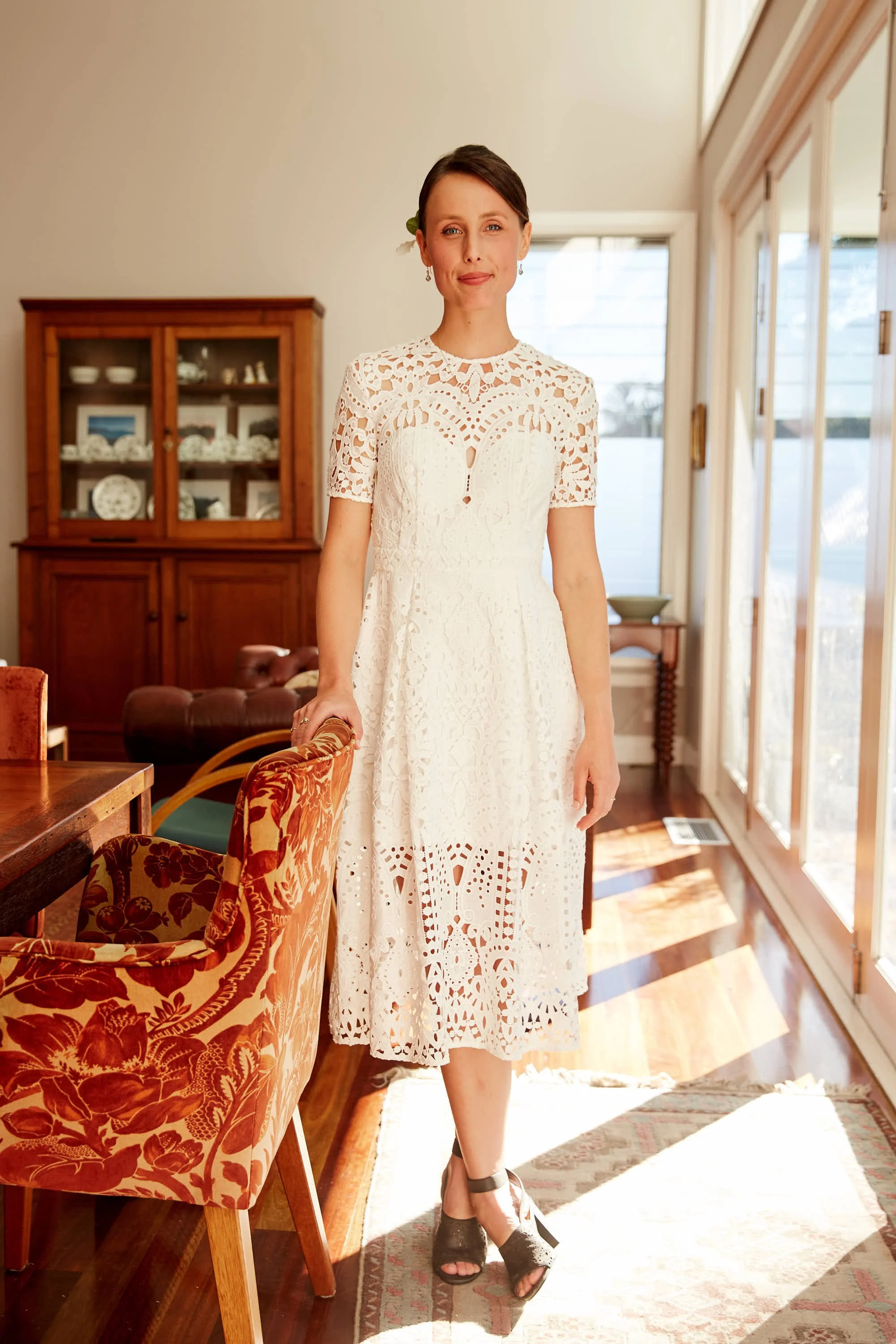 A bride in a white, intricate lace dress standing inside a sunlit room with large windows. She has short, dark hair and is smiling softly.