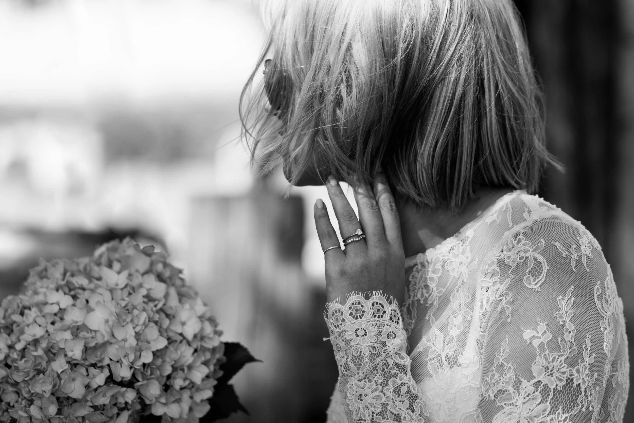 A bride with short blond hair wearing a lace dress standing near a large bouquet of flowers, with her hand gently touching her face.