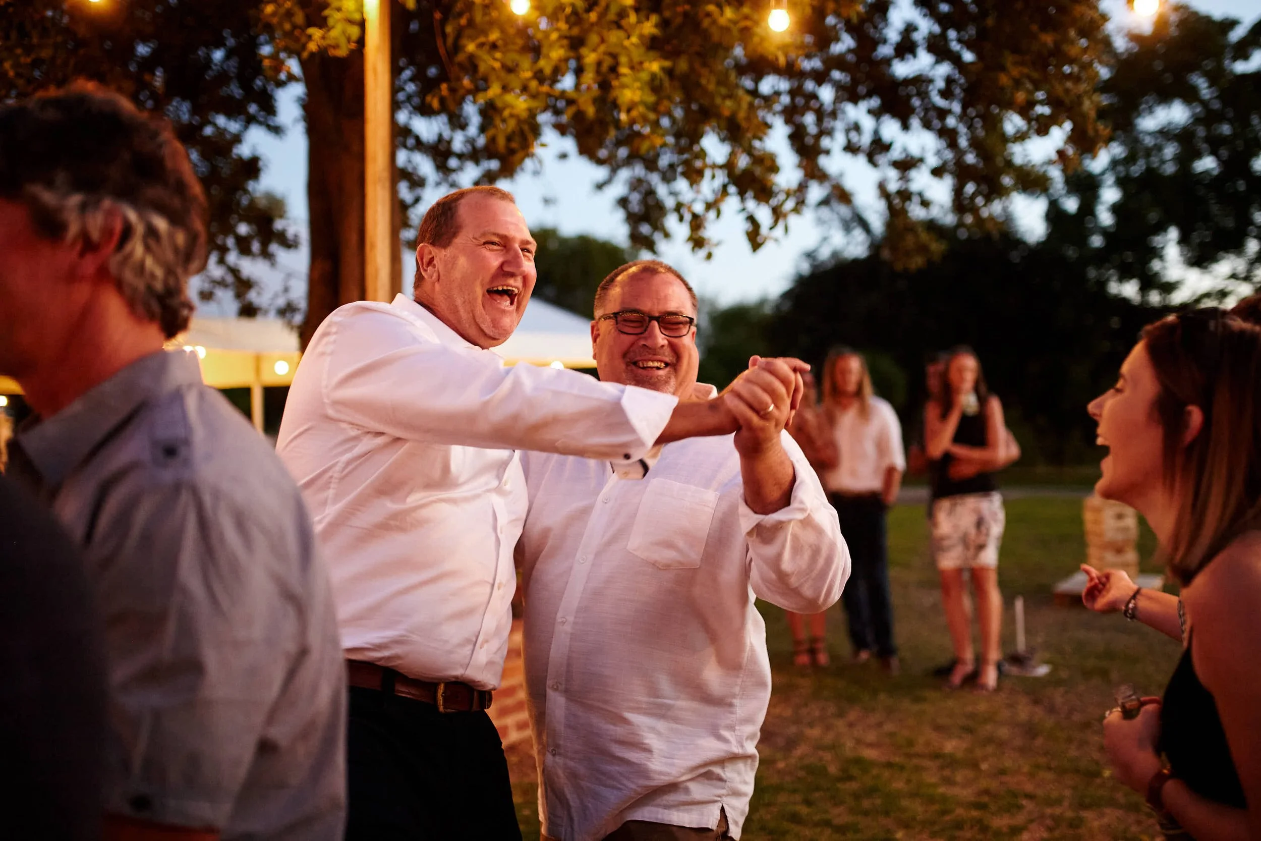 Father and uncle of the bride dancing outdoors at wedding reception, smiling and enjoying themselves, with string lights and trees in the background.