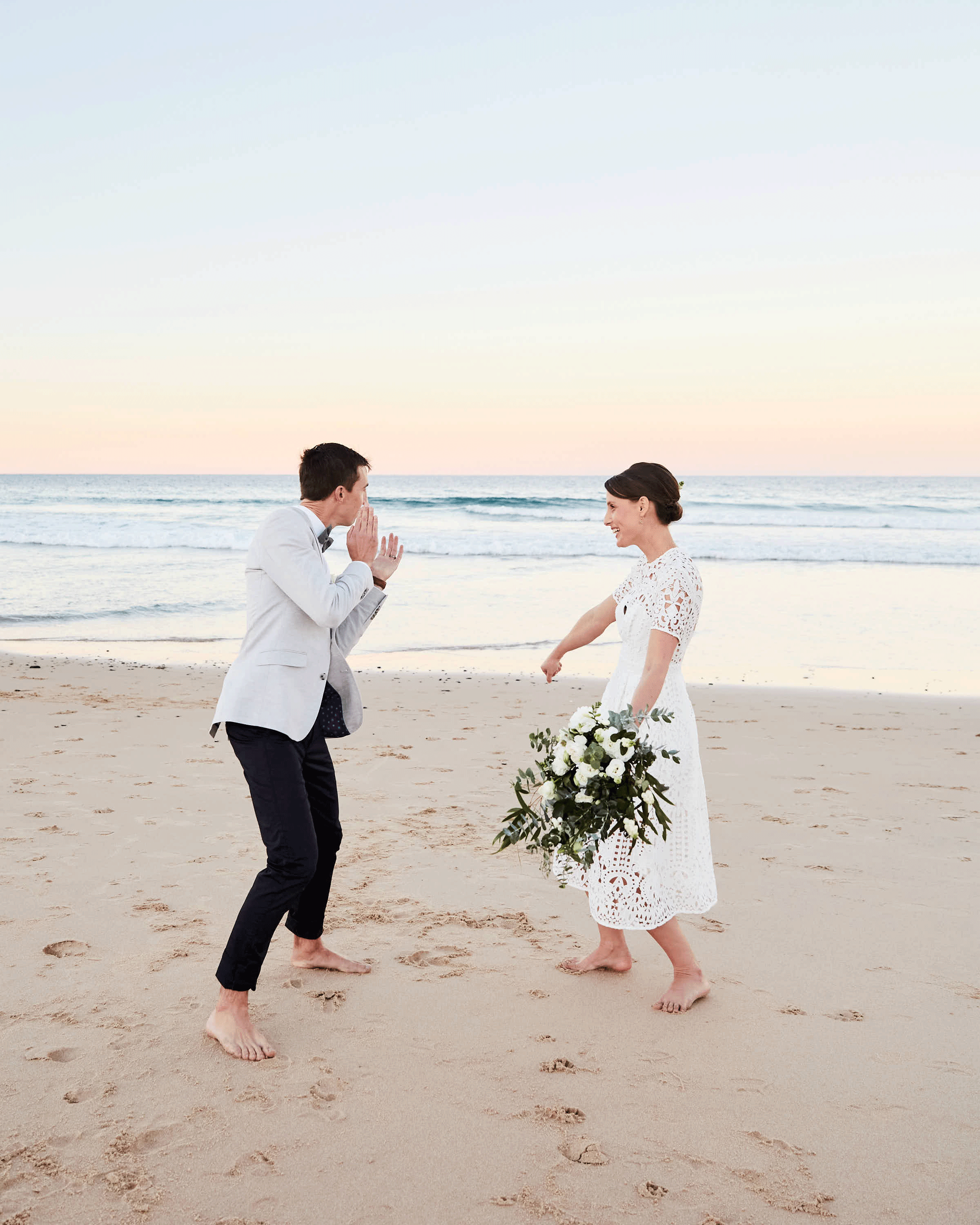 A bride and groom dancing barefoot on the beach during sunset, facing each other, smiling, the bride is holding a bouquet of flowers.
