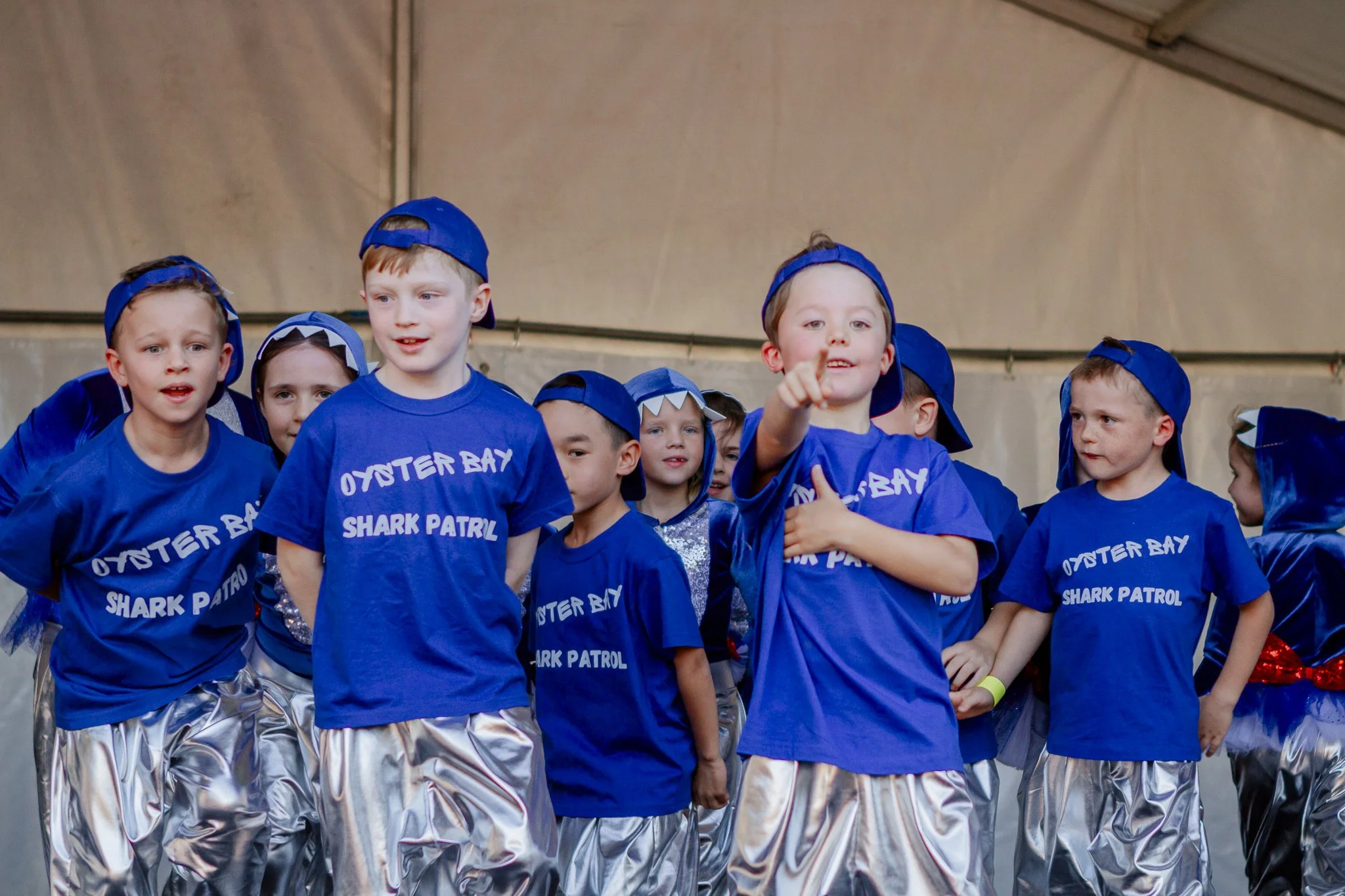 Children dressed in blue shirts and shiny silver pants, performing on stage, some wearing blue caps turned backwards, at an event called 'Oyster Bay Shark Patrol'.