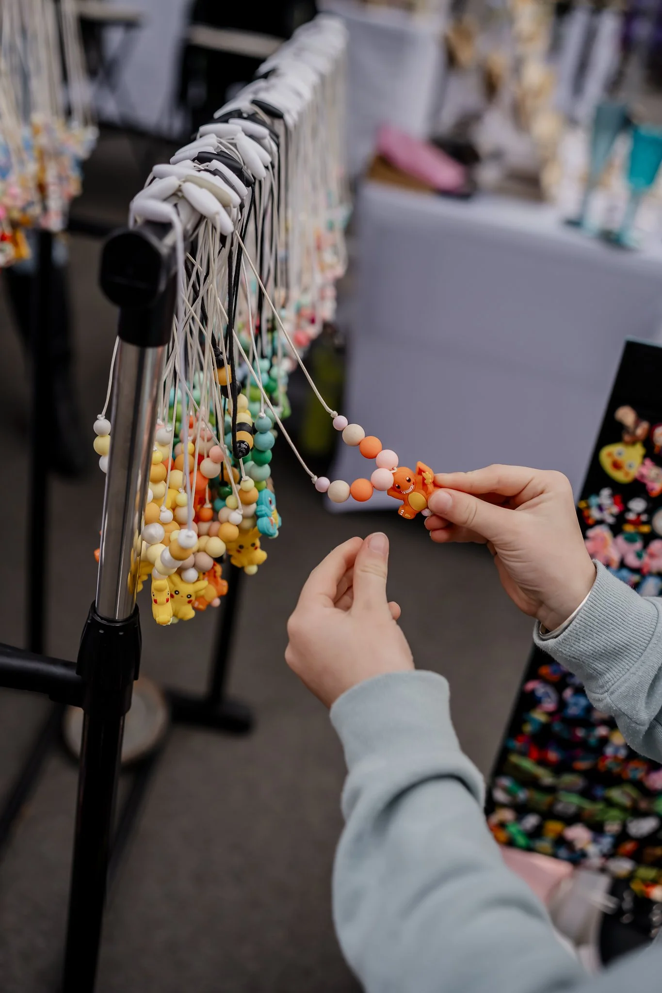 Person holding a beaded necklace with a cute orange creature pendant at a jewelry stall with similar necklaces hanging on display at the Oyster Bay Arts and Craft Festival.