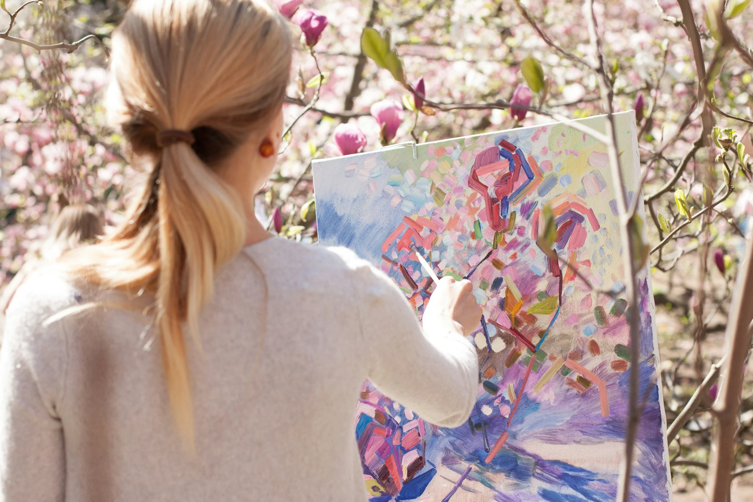 A woman with blonde hair painting on a canvas outdoors surrounded by blooming pink magnolia flowers.