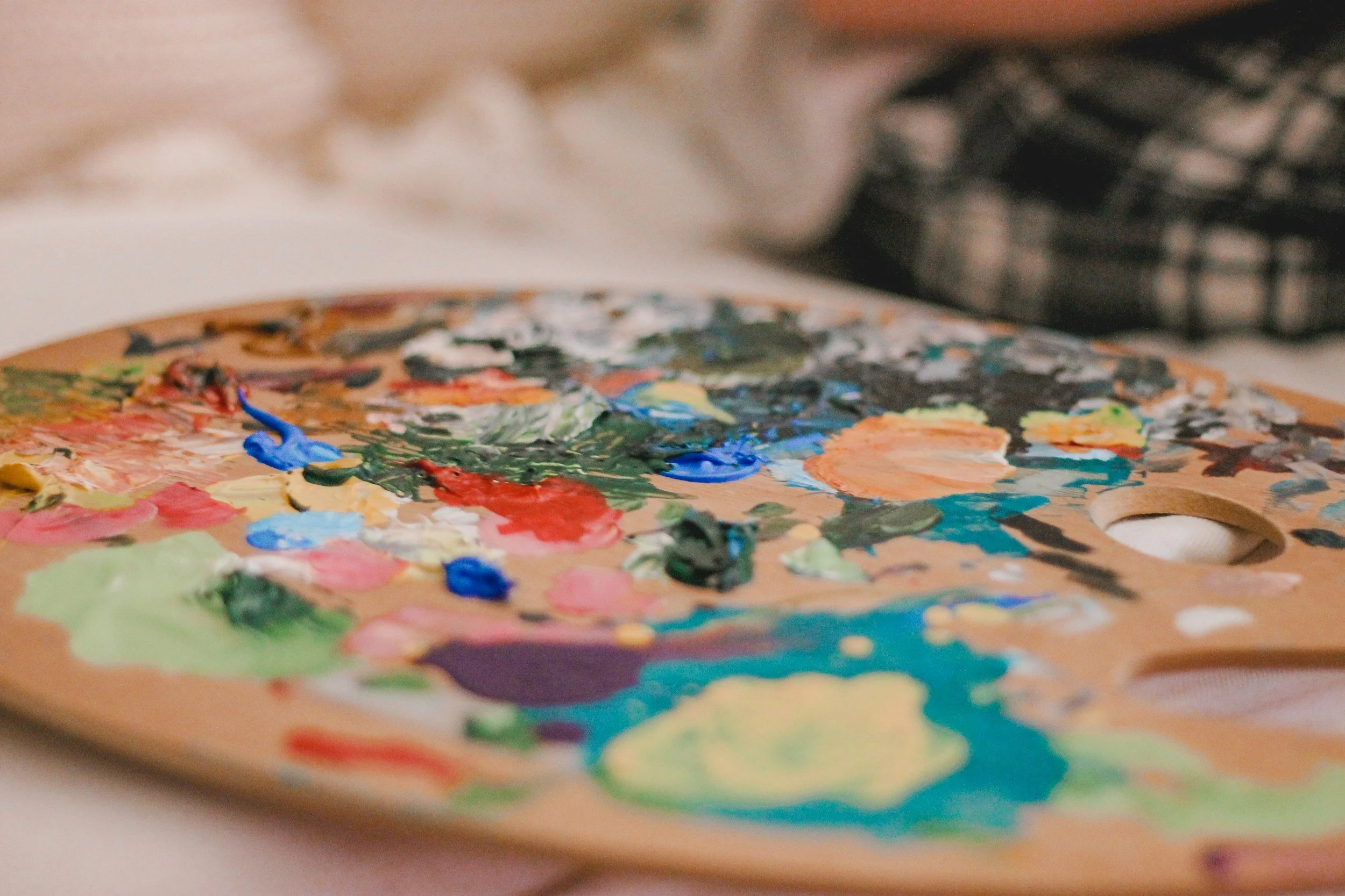 A wooden artist's palette with various blobs of colorful paint, including blue, green, red, yellow, and orange, resting on a surface with a blurred background.