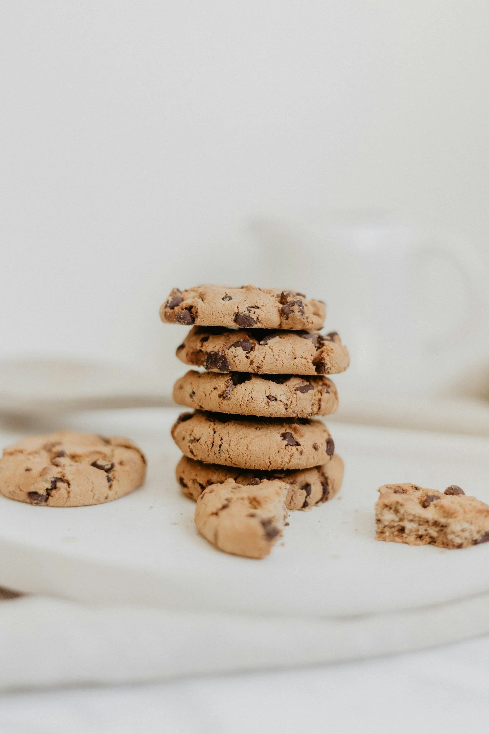 Stack of five chocolate chip cookies on a white surface with a few broken cookie pieces nearby.