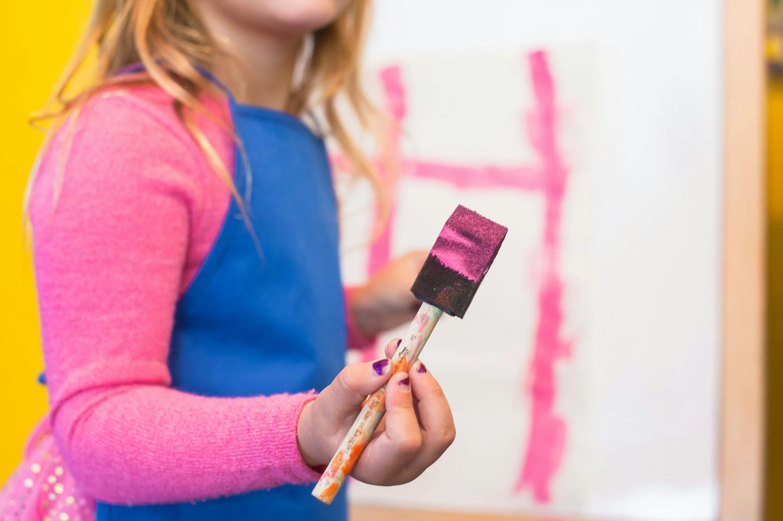 A girl in a pink long-sleeve shirt and blue apron holding a paintbrush with pink and black paint, standing in front of a white canvas with pink streaks at the Oyster Bay Art and Craft Festival.