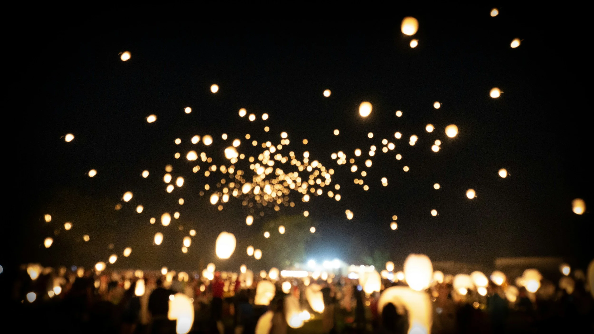 Nighttime scene of sky lanterns floating upward during a festival or event, with many lanterns lit and rising into the dark sky, and a crowd of people below watching.