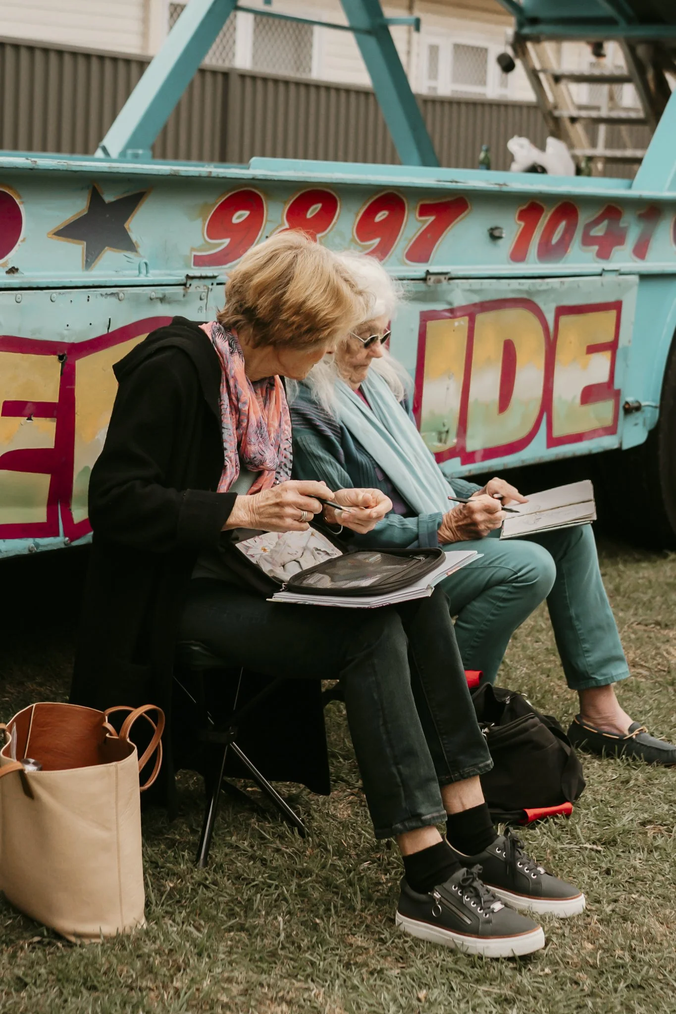 Artist sitting outdoors next to a colorful carnival ride, looking at notebooks or clipboards at the Oyster Bay Arts and Craft Festival.