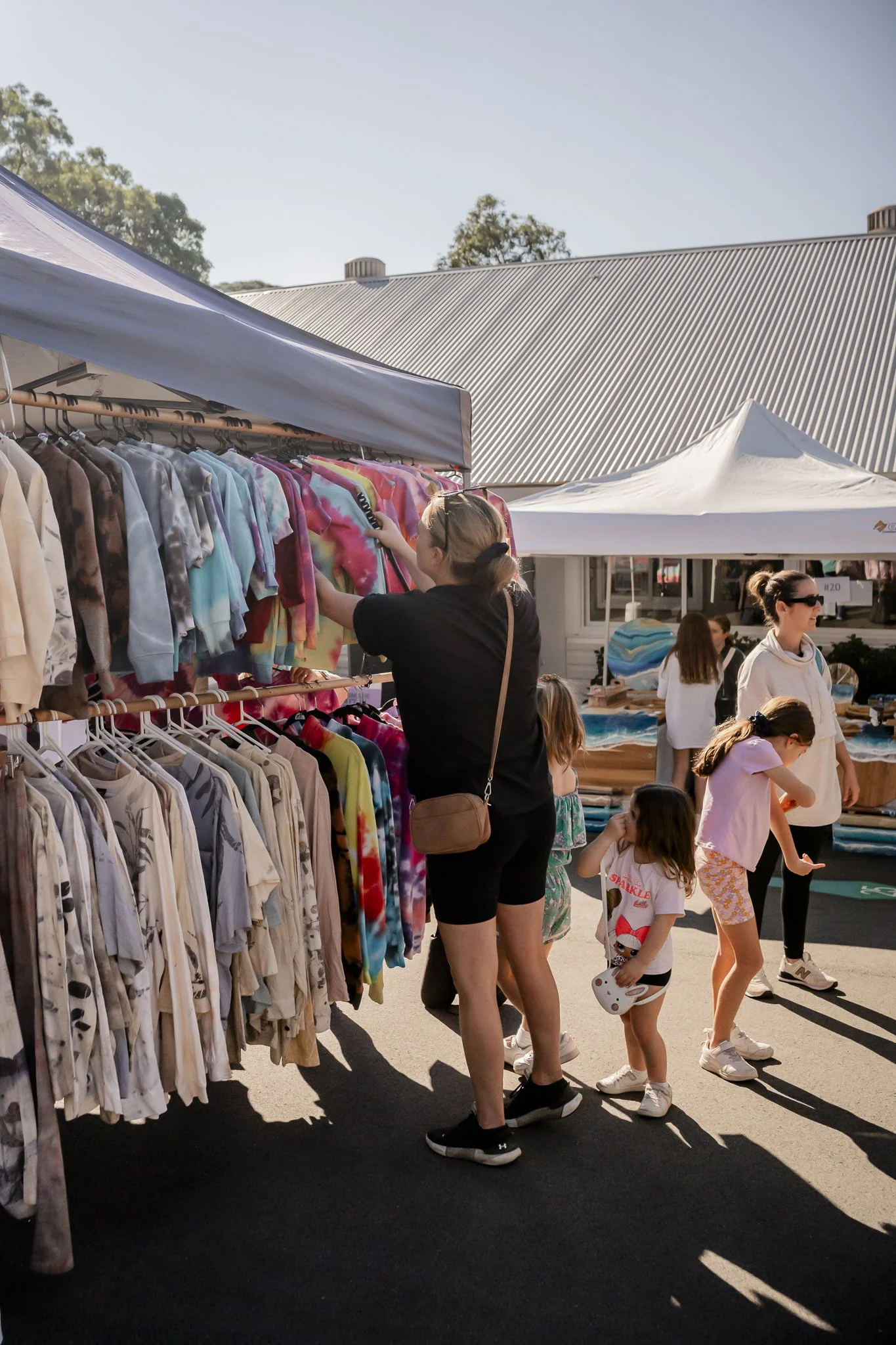 People shopping at an outdoor market with clothing and art displays, some children and women looking at items.