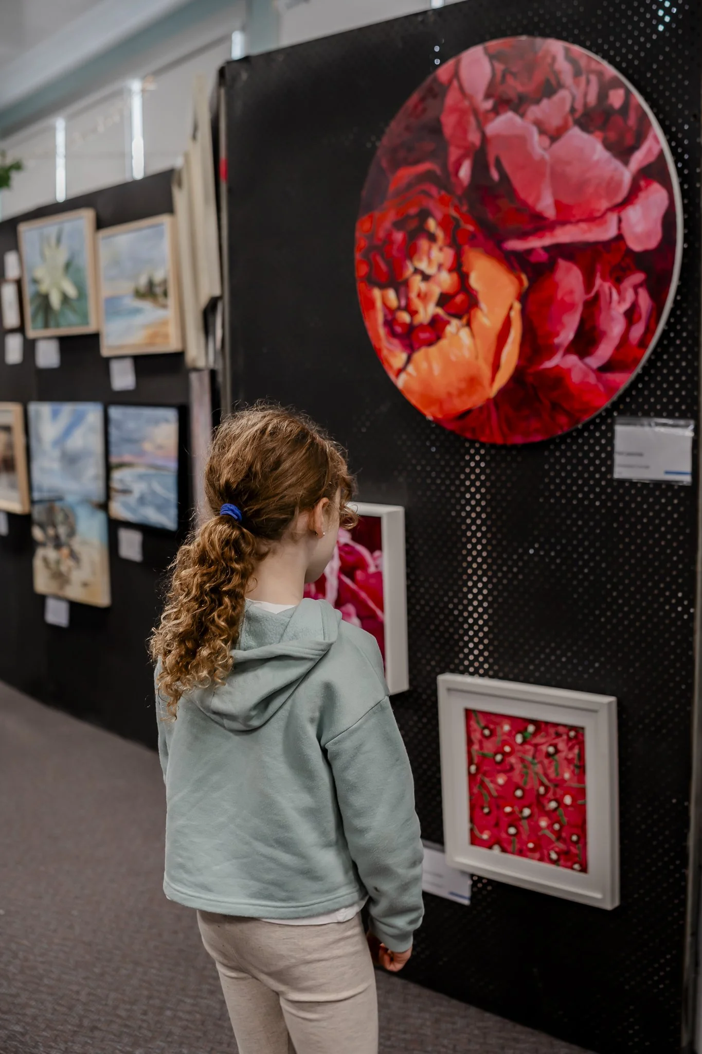 A young girl with curly hair tied back in a ponytail, wearing a light grey hoodie and beige pants, is observing artwork at an art gallery. The artwork includes a large circular painting of assorted pink and red rocks and smaller framed paintings of f